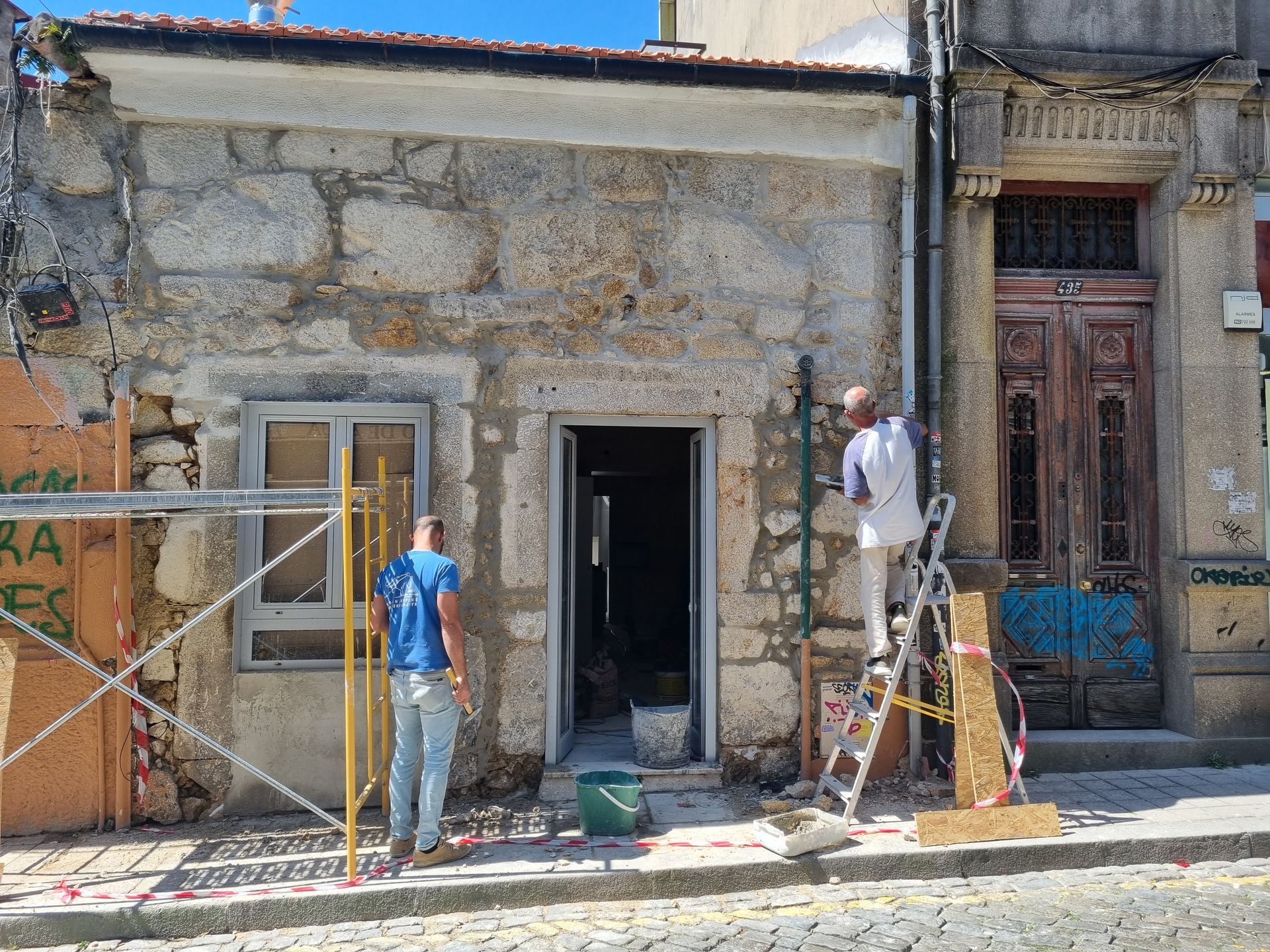 Deux personnes travaillent à la restauration de la façade en pierre d'un bâtiment, à l'aide d'échafaudages et d'une échelle, dans une rue de la ville.