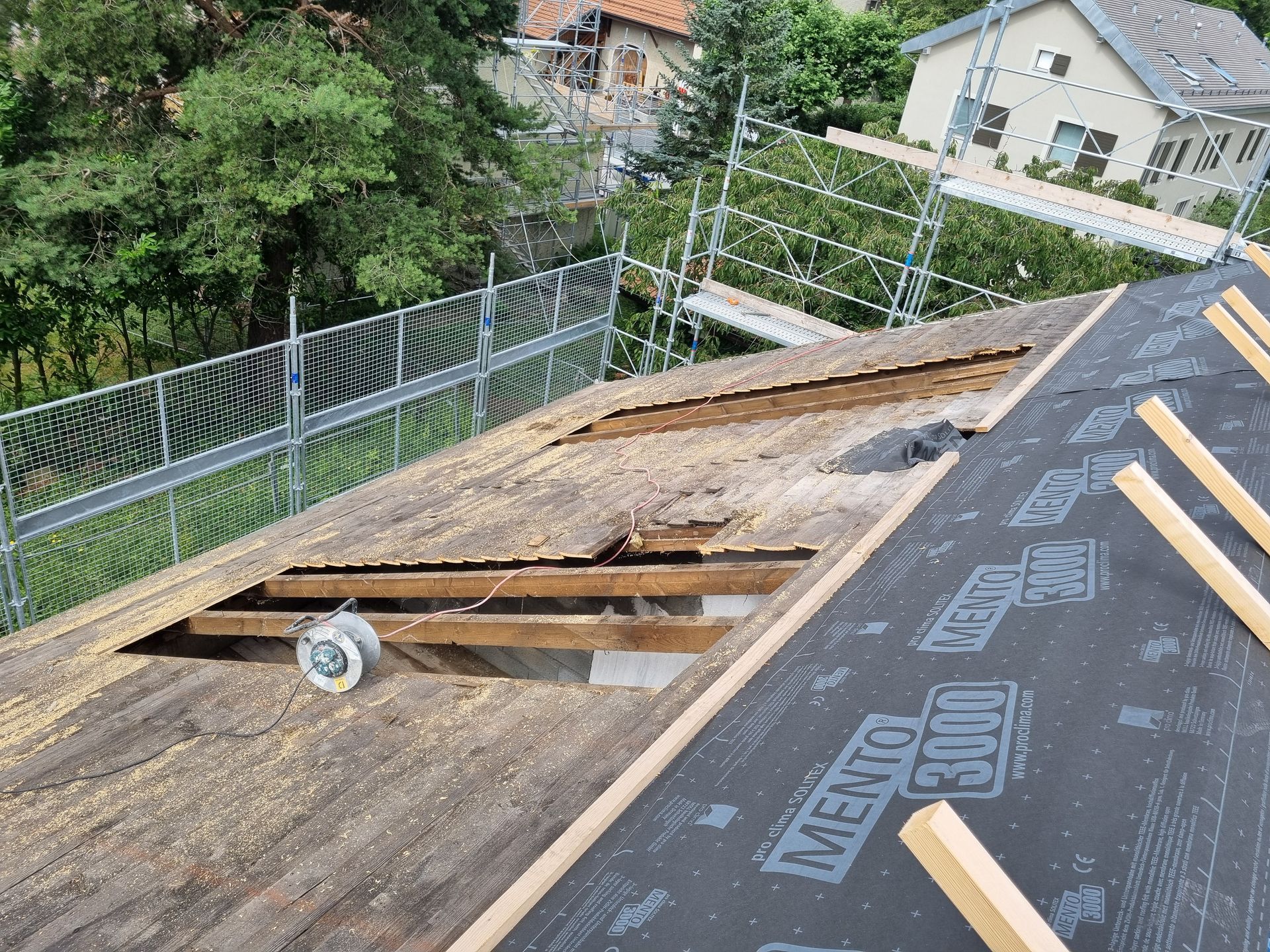 Toiture en rénovation, laissant apparaître les chevrons en bois, une section recouverte d'une membrane noire et les échafaudages environnants.