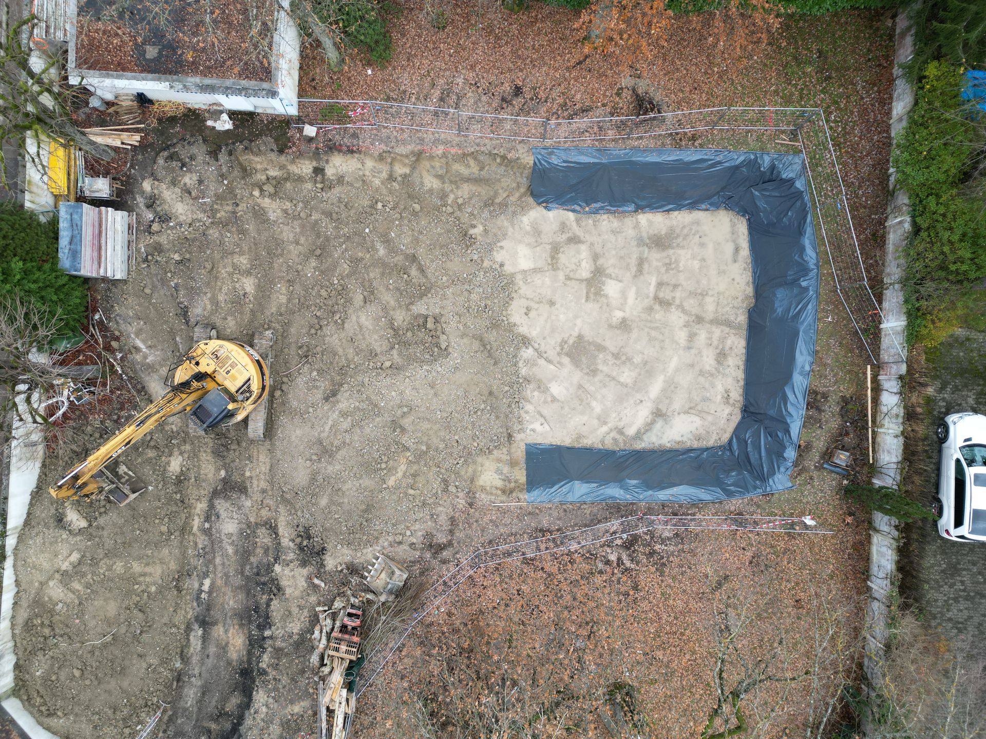 Vue aérienne d'un chantier de construction montrant une excavatrice jaune à côté d'une zone excavée recouverte de plastique noir.