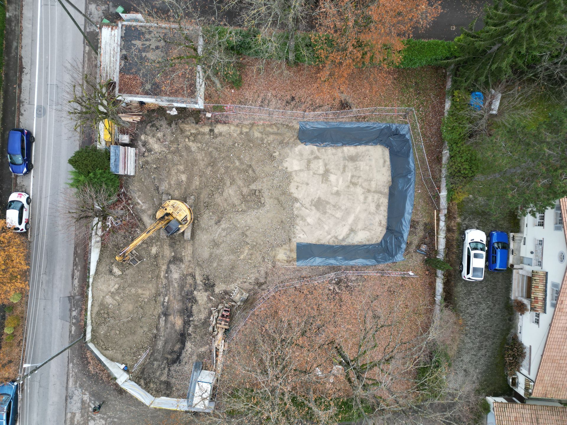 Vue aérienne d'un chantier de construction avec une excavatrice, une fondation en sable préparée et des propriétés résidentielles environnantes.