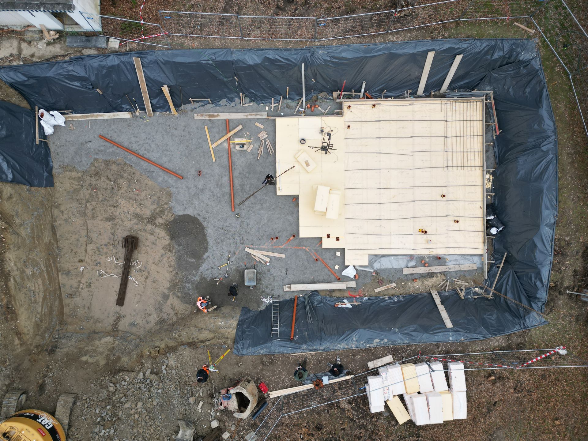 Vue aérienne des fondations d'un chantier de construction avec des panneaux de bois, du gravier et des ouvriers à l'intérieur d'une fosse clôturée.