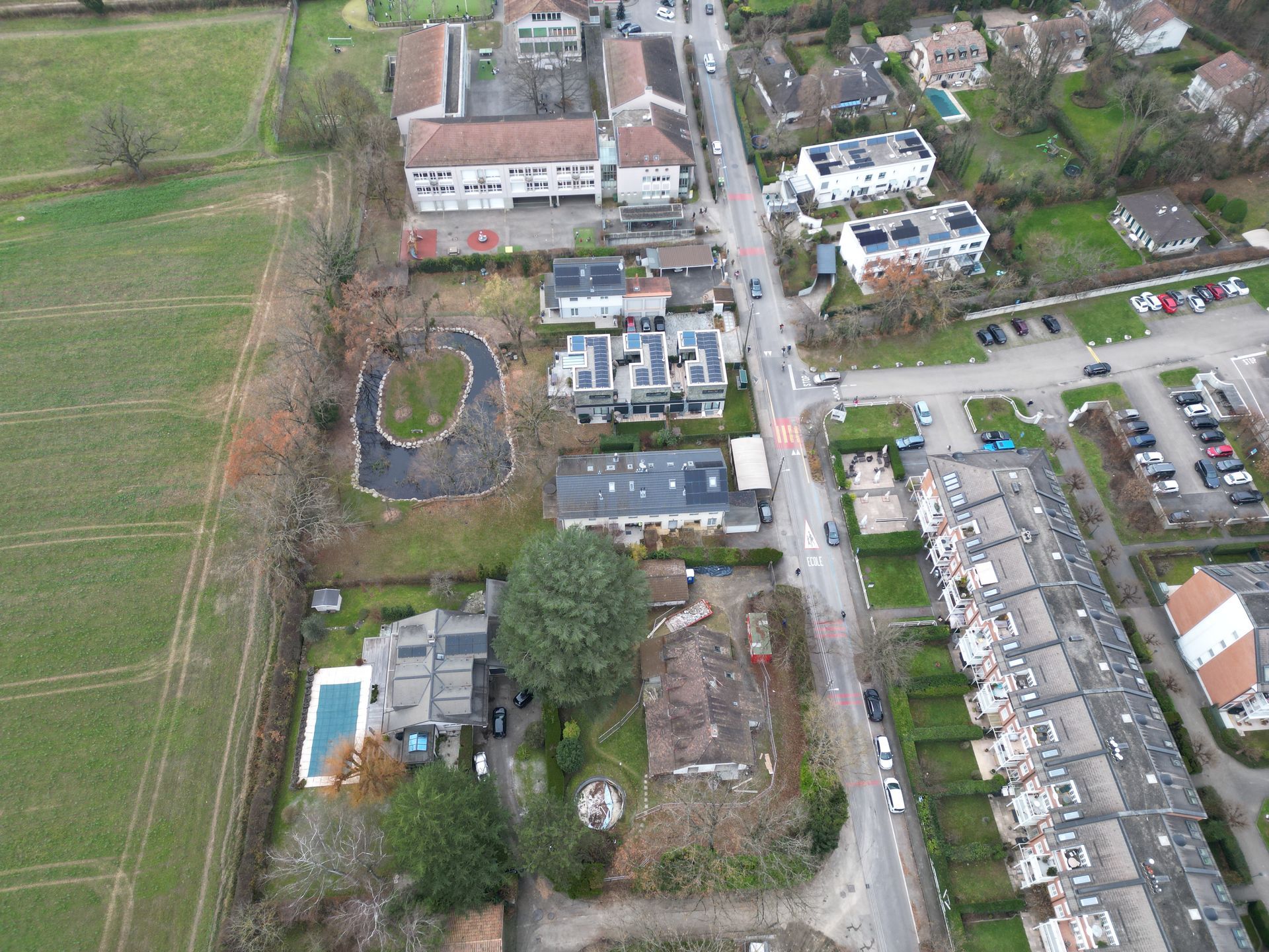 Vue aérienne d'un quartier résidentiel avec des maisons, une piscine, un petit étang et un champ.