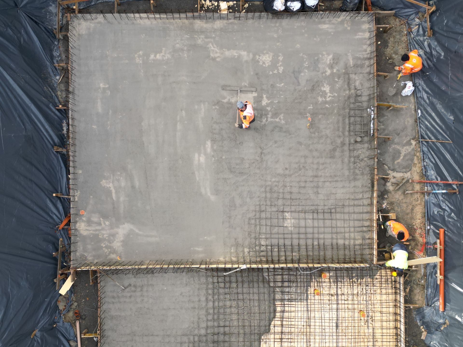Vue aérienne d'ouvriers du bâtiment coulant du béton dans une dalle de fondation à ossature métallique sur un chantier recouvert de plastique noir.