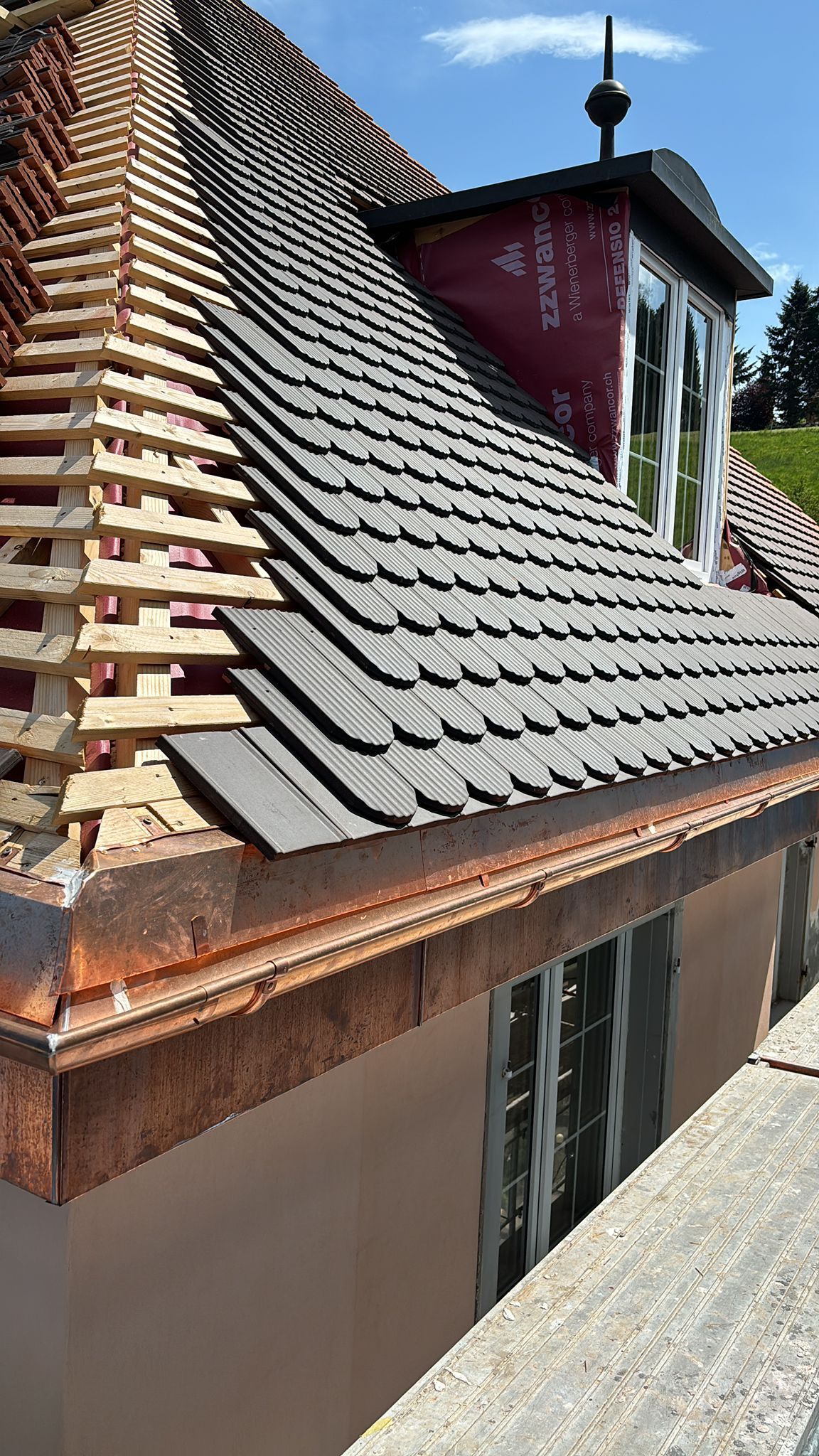 Vue latérale d'un toit de maison en construction, avec des tuiles gris foncé partiellement posées et un lattis de bois visible.