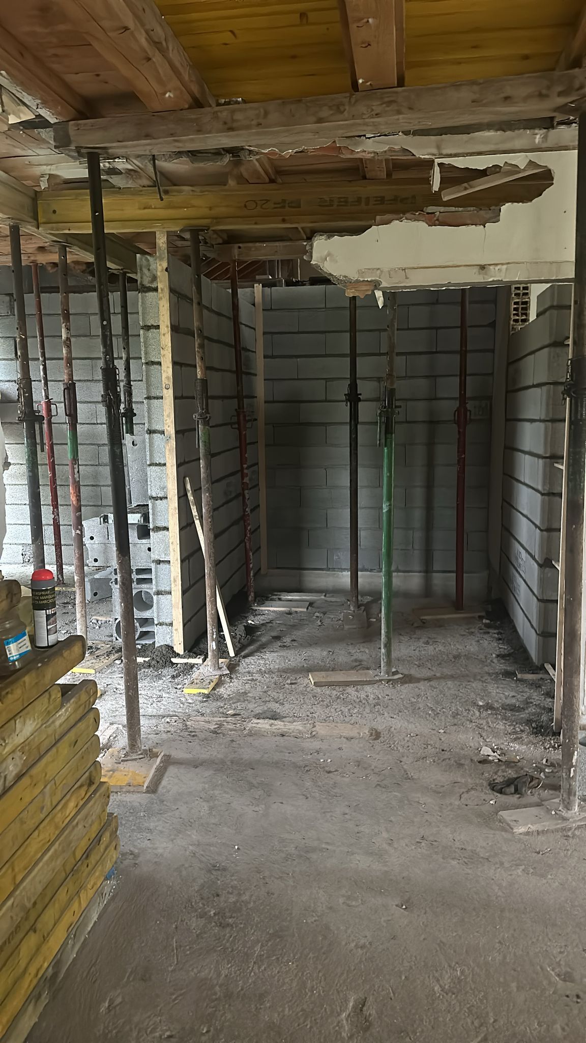 Un chantier de construction intérieur montrant des supports en acier temporaires soutenant les solives de plafond au-dessus de murs en blocs de béton brut.