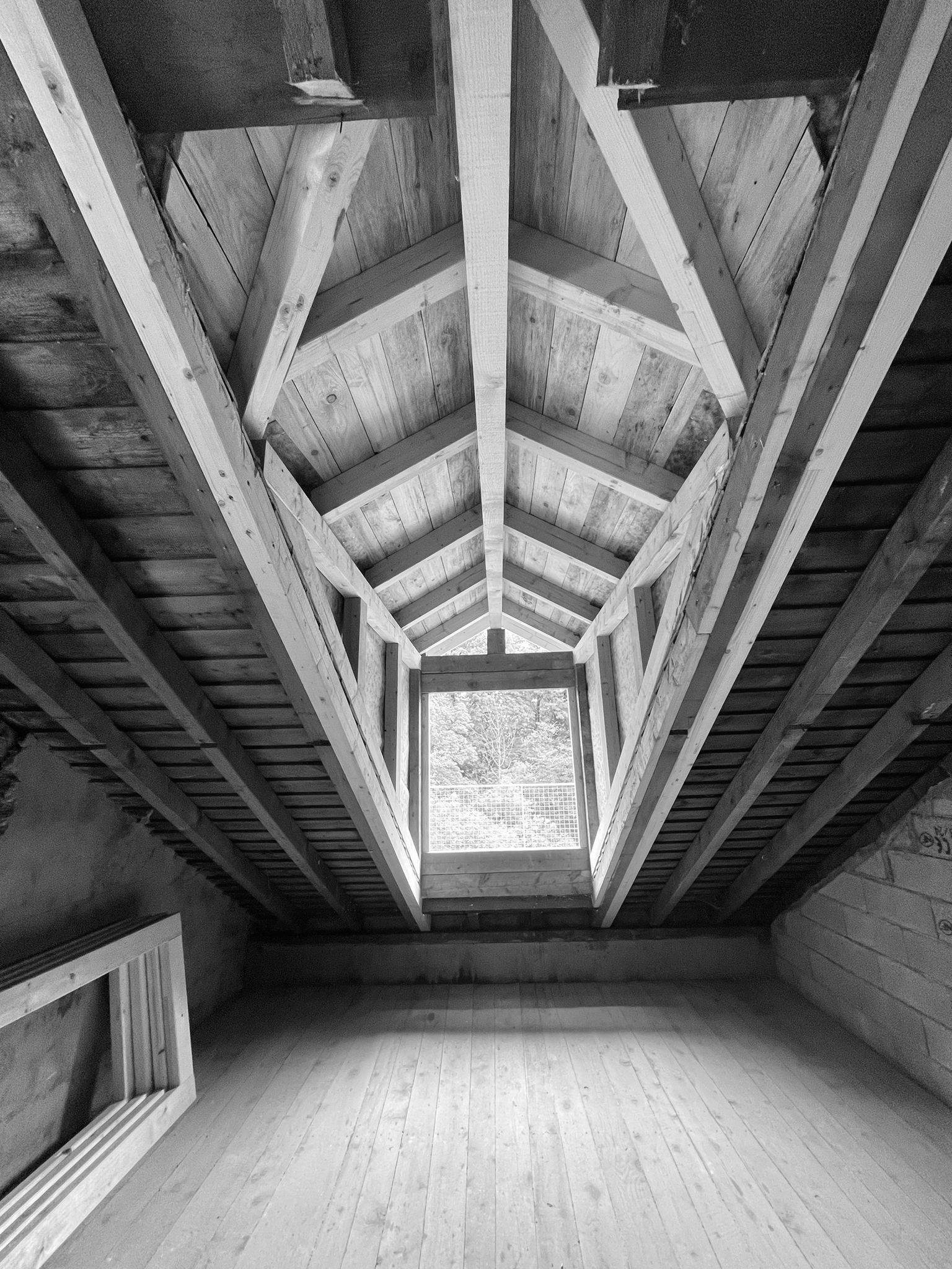 Vue en noir et blanc de l'intérieur d'un grenier avec des poutres apparentes, un plancher en bois clair et une fenêtre encadrée.