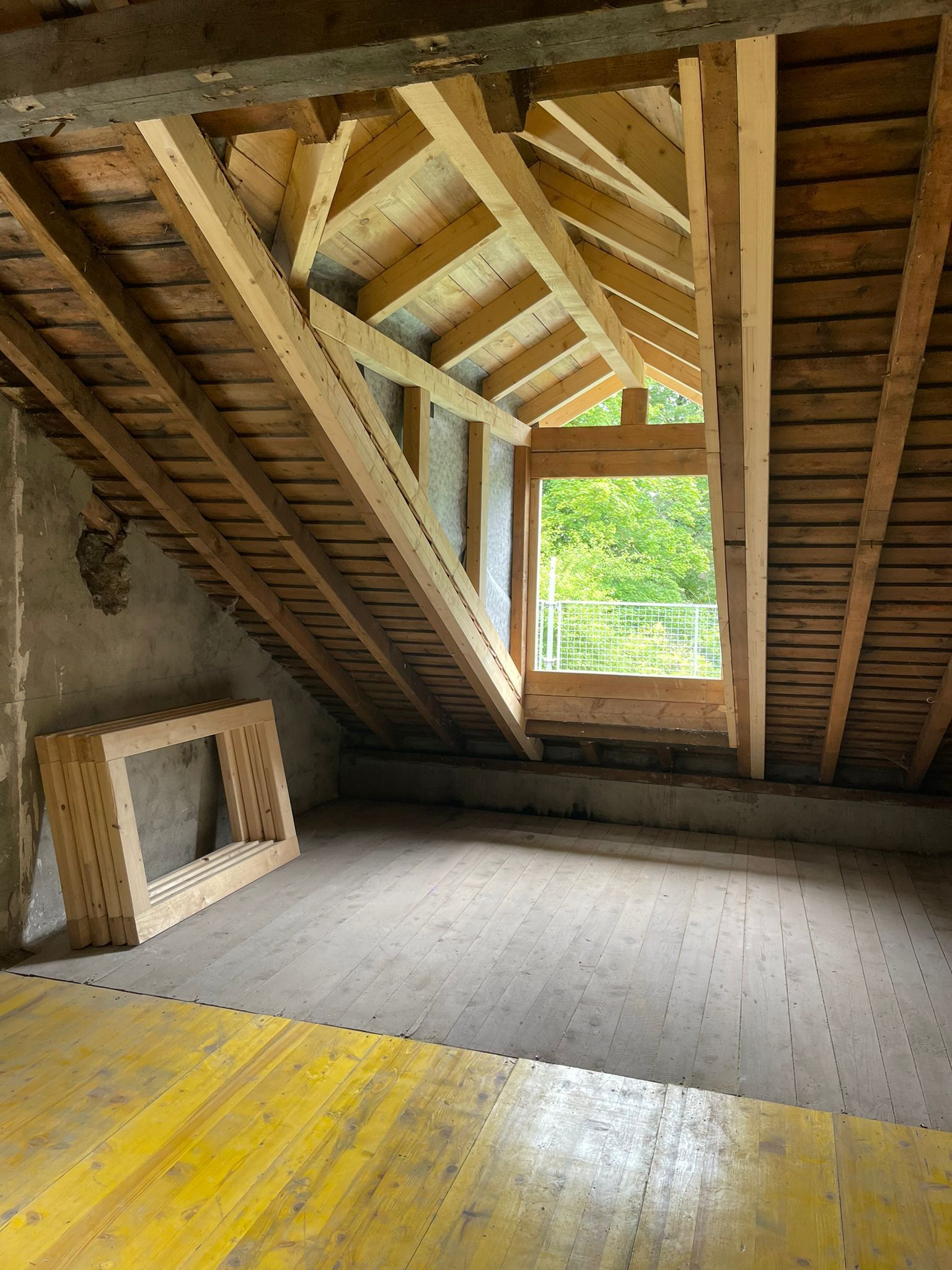 Grenier en cours de rénovation avec poutres apparentes, nouveau cadre de fenêtre et panneaux de sol jaunes.