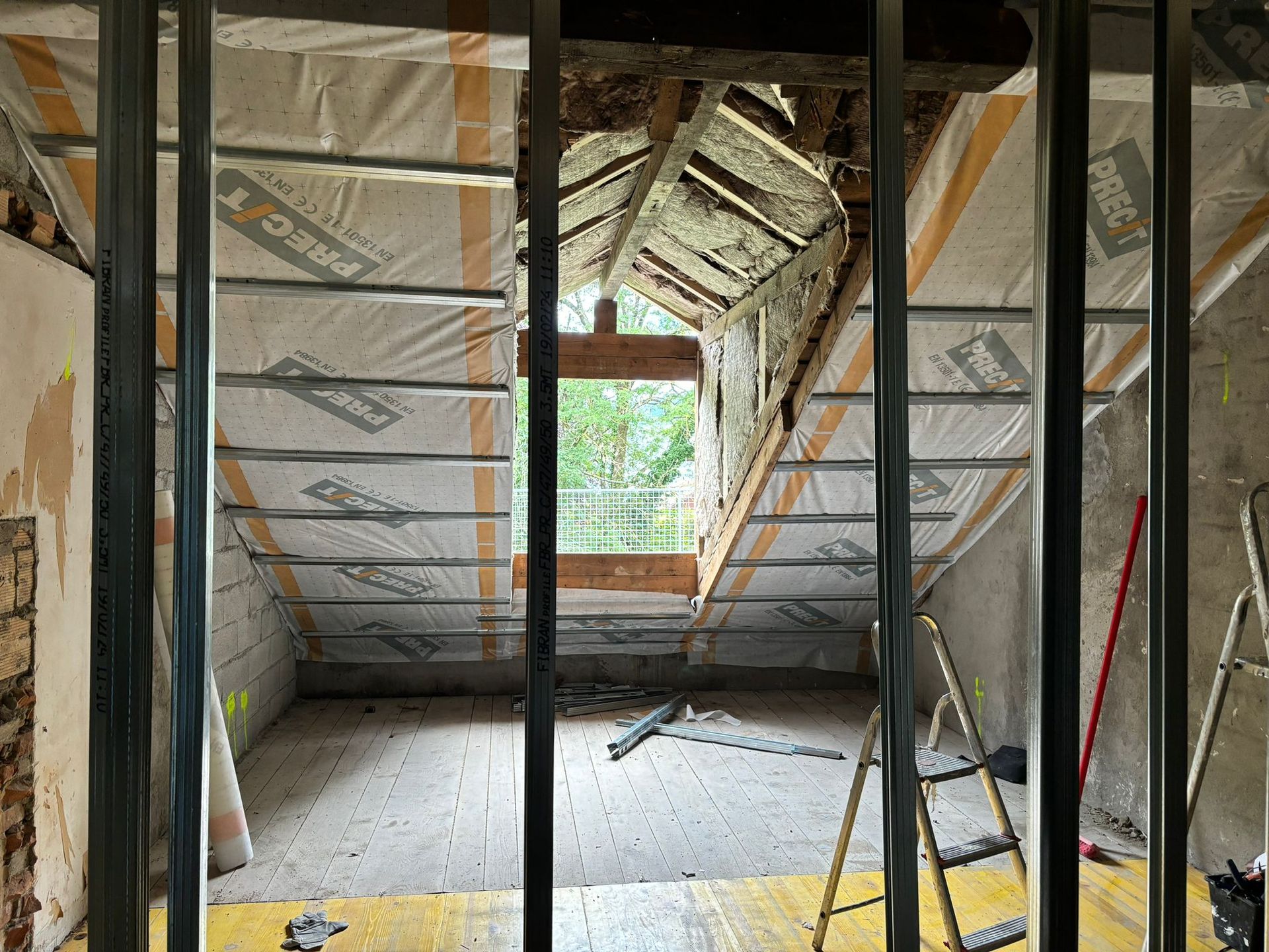 Un grenier en cours de rénovation avec plafond incliné isolé, charpente métallique, une fenêtre centrale et un escabeau.