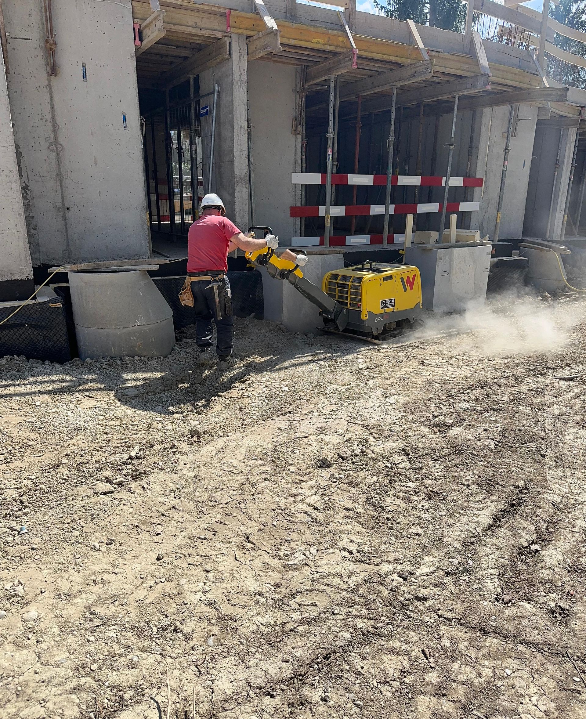 Un ouvrier du bâtiment en chemise rouge utilise une plaque vibrante jaune pour niveler la terre à côté d'un bâtiment en béton inachevé.