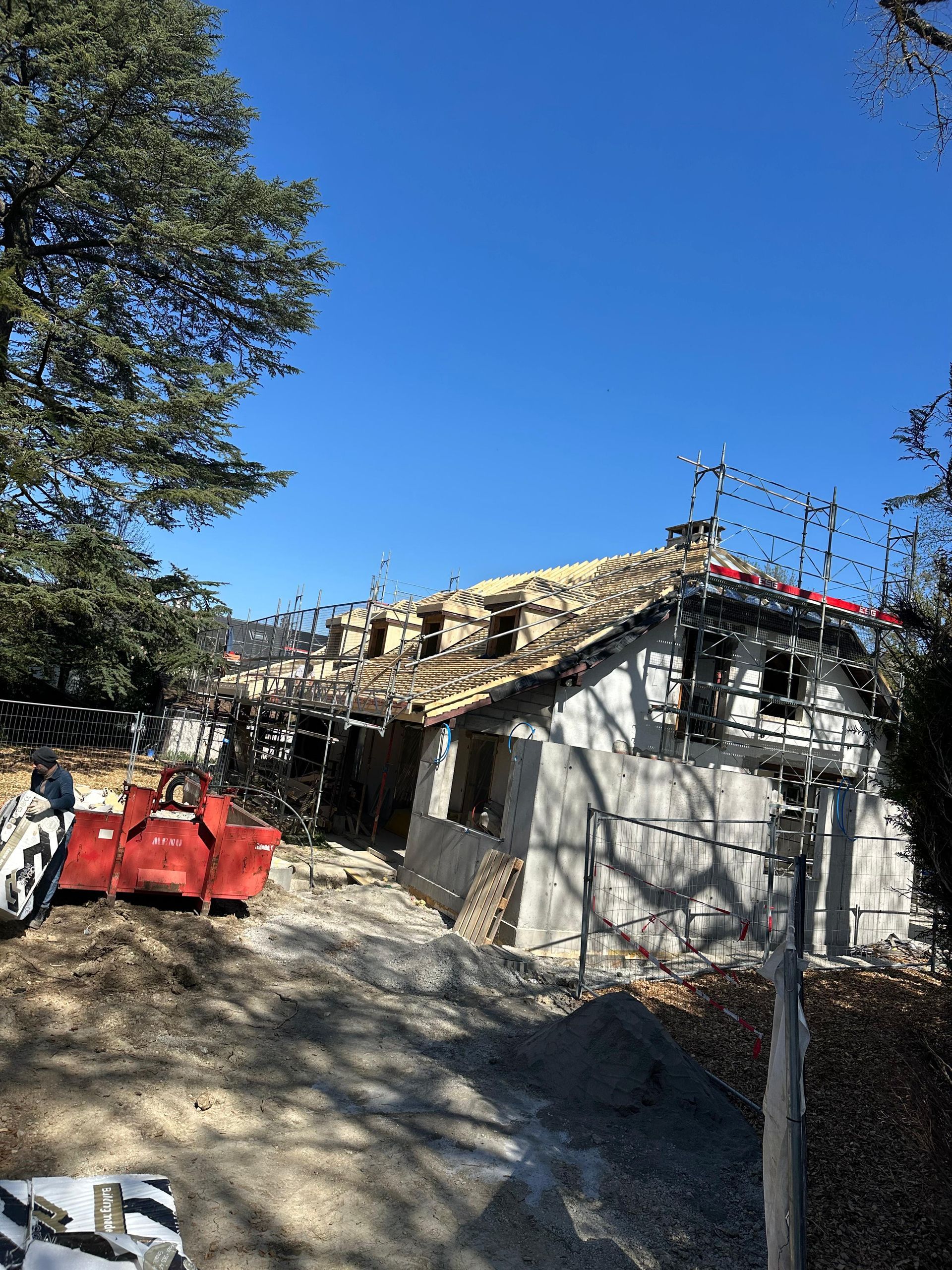 Une maison en construction avec des échafaudages sur le toit et un conteneur à gravats rouge sur le terrain vague, sous un ciel bleu.