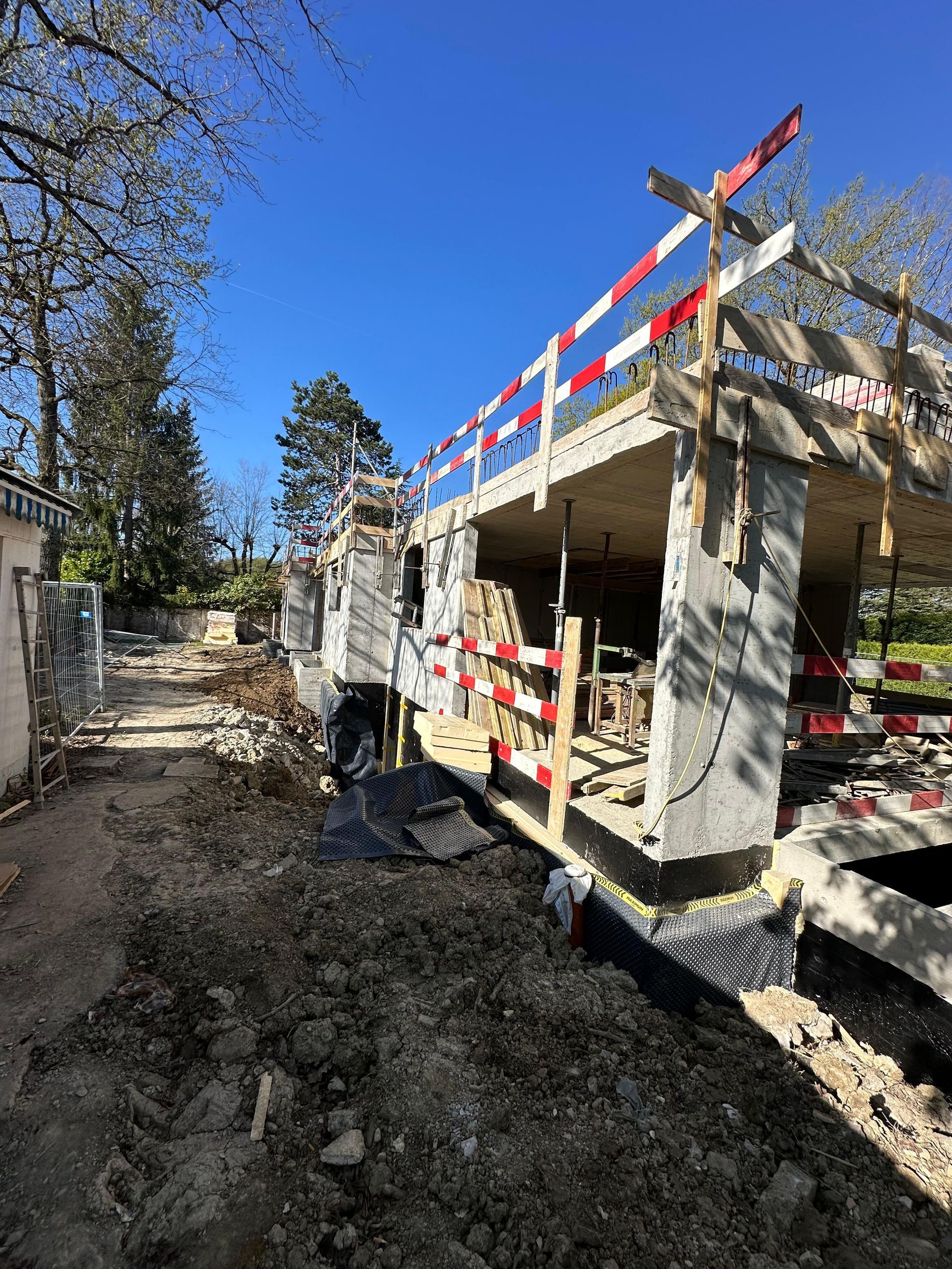 Un chantier de construction montrant une structure en béton avec des barrières de sécurité en ruban adhésif rouge et blanc sous un ciel bleu dégagé.