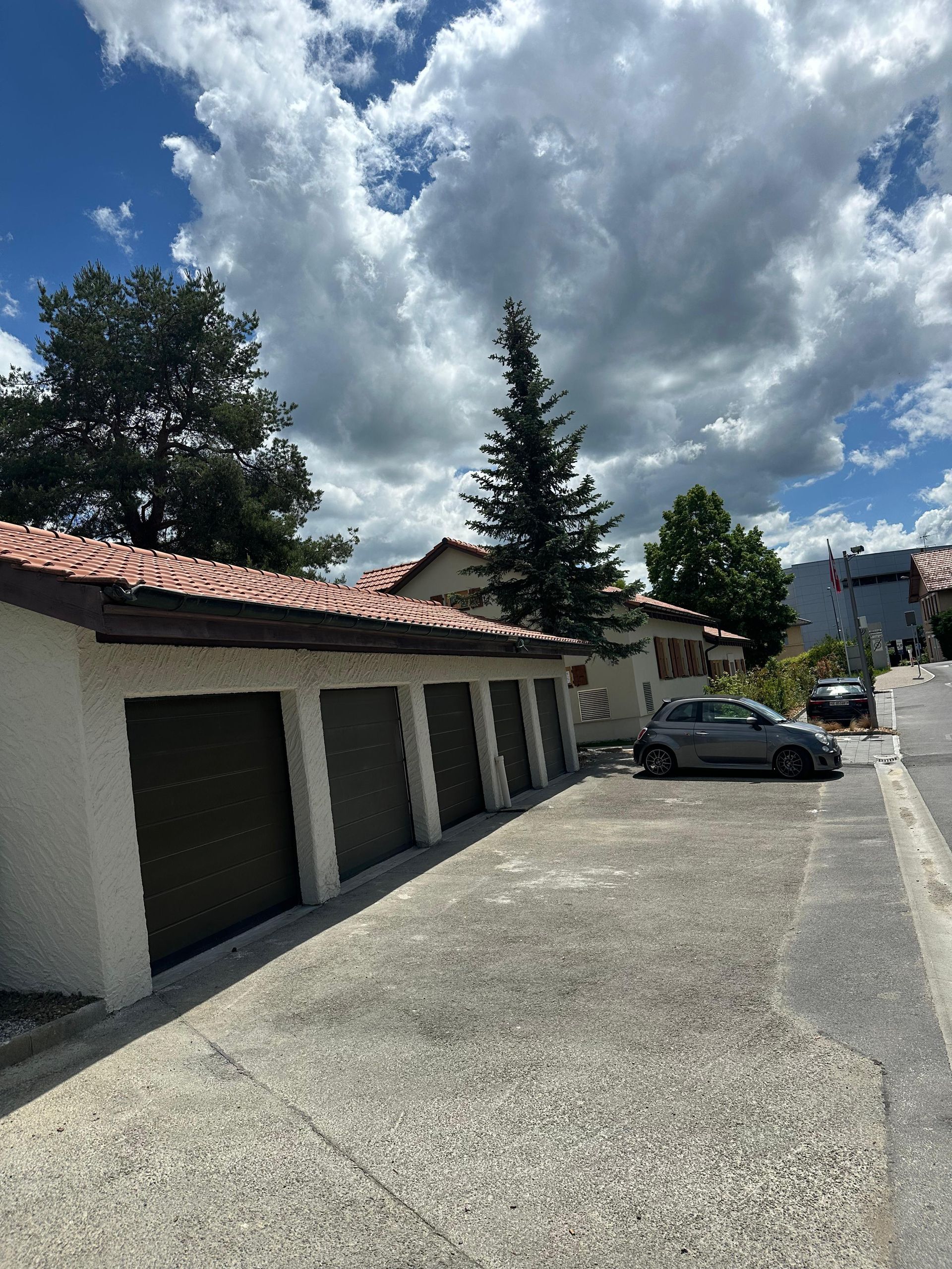 Une rangée de garages aux murs blancs et aux toits de tuiles rouges, avec une voiture grise garée à côté sous un ciel lumineux et nuageux.