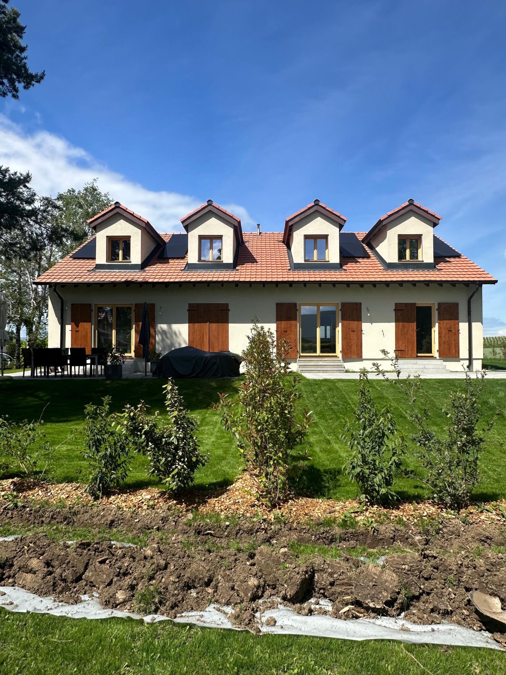 Une maison à deux étages avec un toit en tuiles de terre cuite, quatre lucarnes et des volets en bois, se détachant sur une pelouse et un ciel bleu.