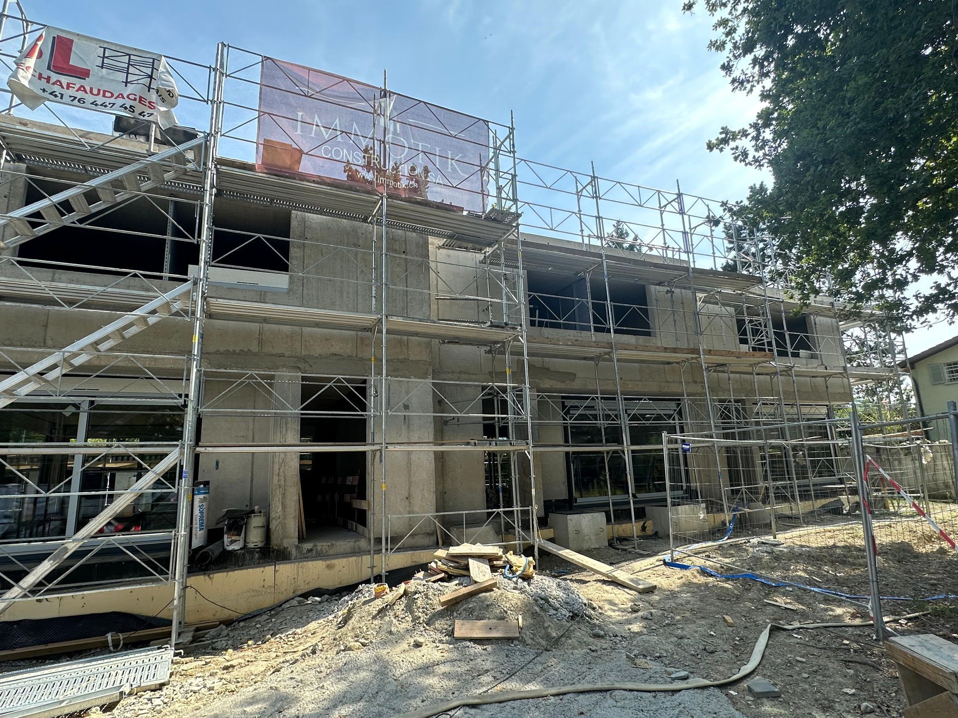 Un bâtiment de deux étages en construction, recouvert d'échafaudages métalliques, avec une enseigne fixée à l'étage supérieur.