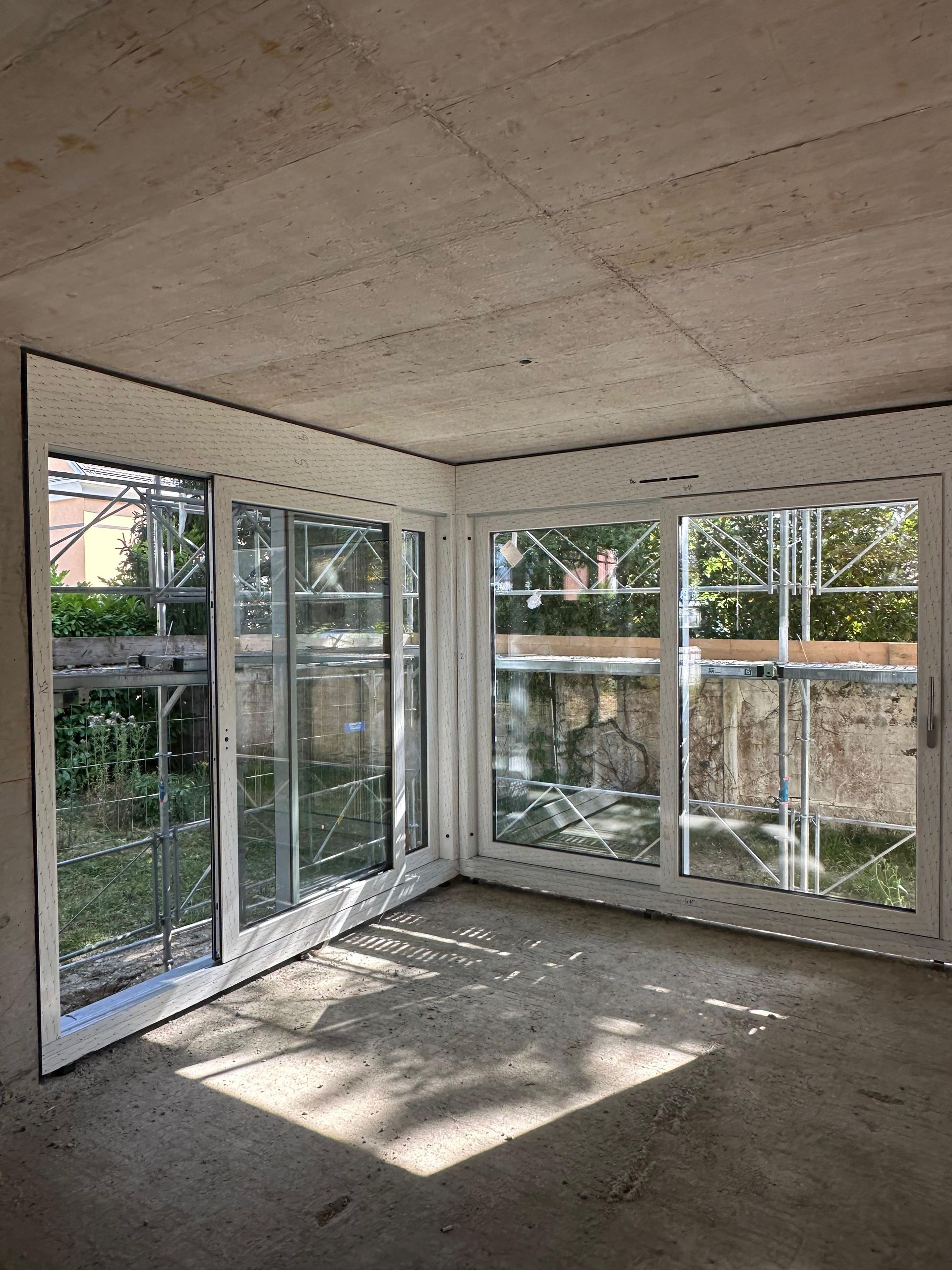 Coin d'une pièce en construction avec de grandes fenêtres vitrées, des cadres blancs et des murs et un sol en béton brut.