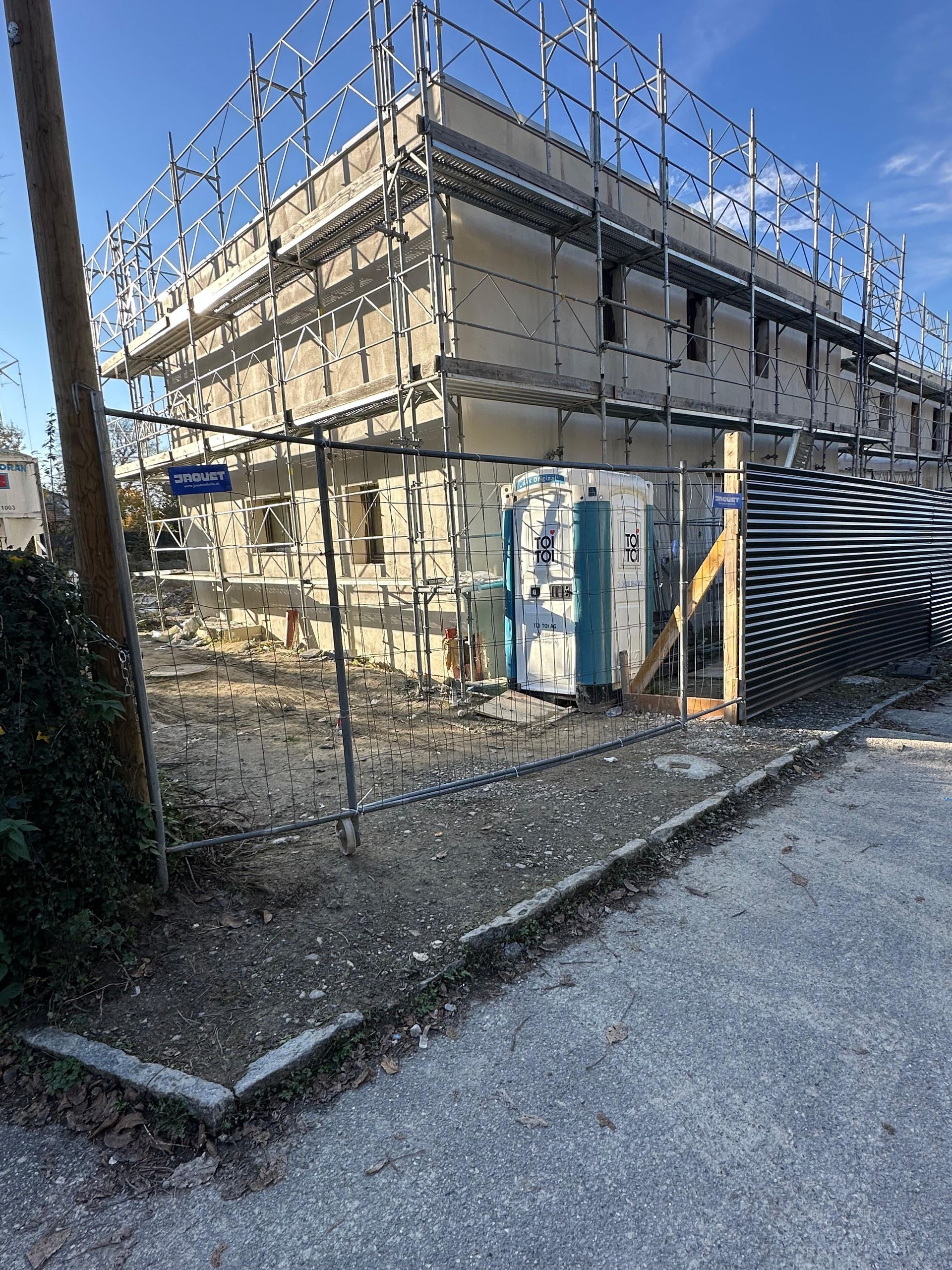 Un bâtiment de deux étages en construction, entouré d'échafaudages et d'une clôture en fil de fer, avec des toilettes portables dans la cour.