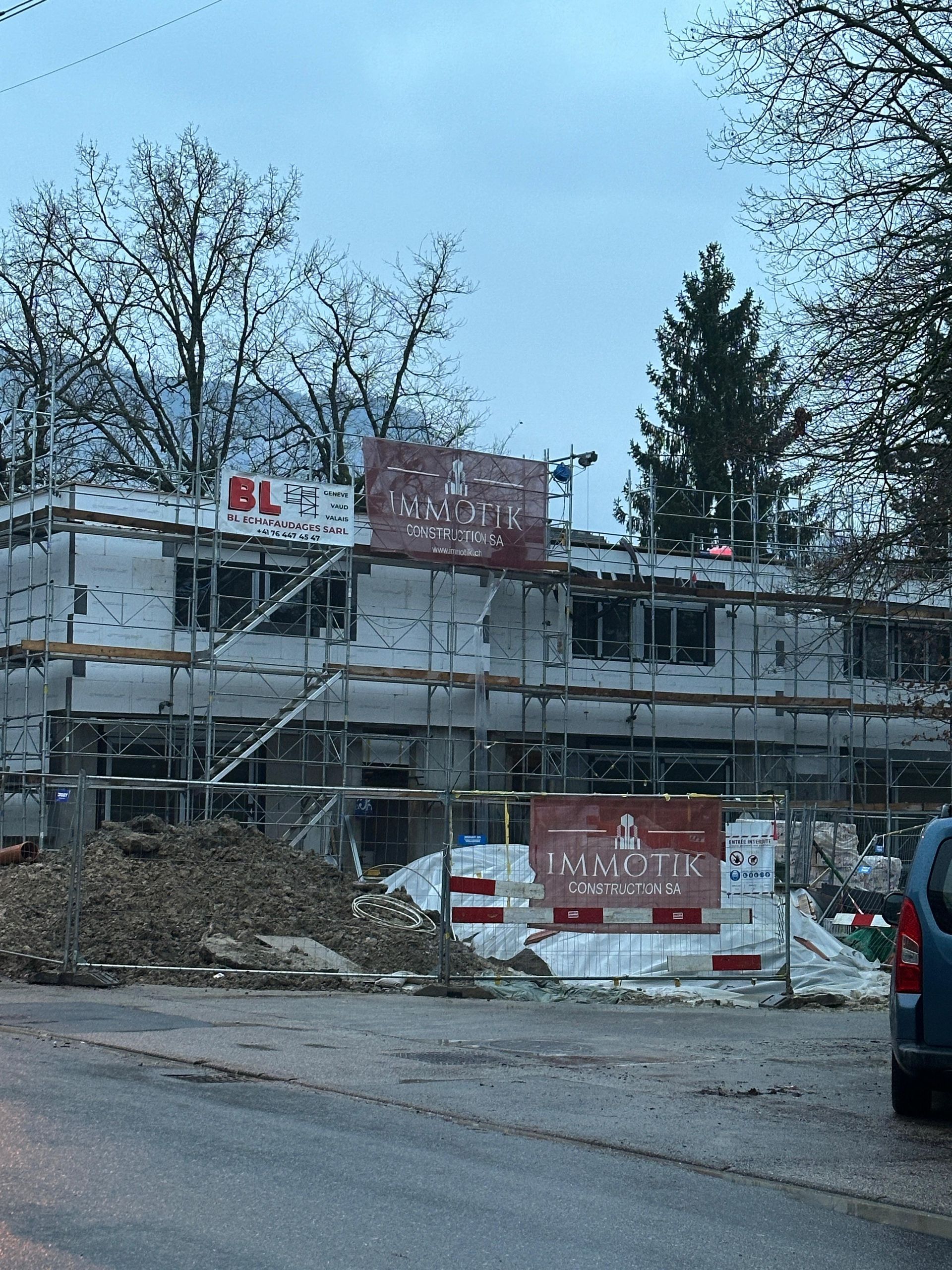 Un bâtiment en construction, recouvert de panneaux isolants blancs, avec des échafaudages et des panneaux de signalisation, dans un cadre boisé.