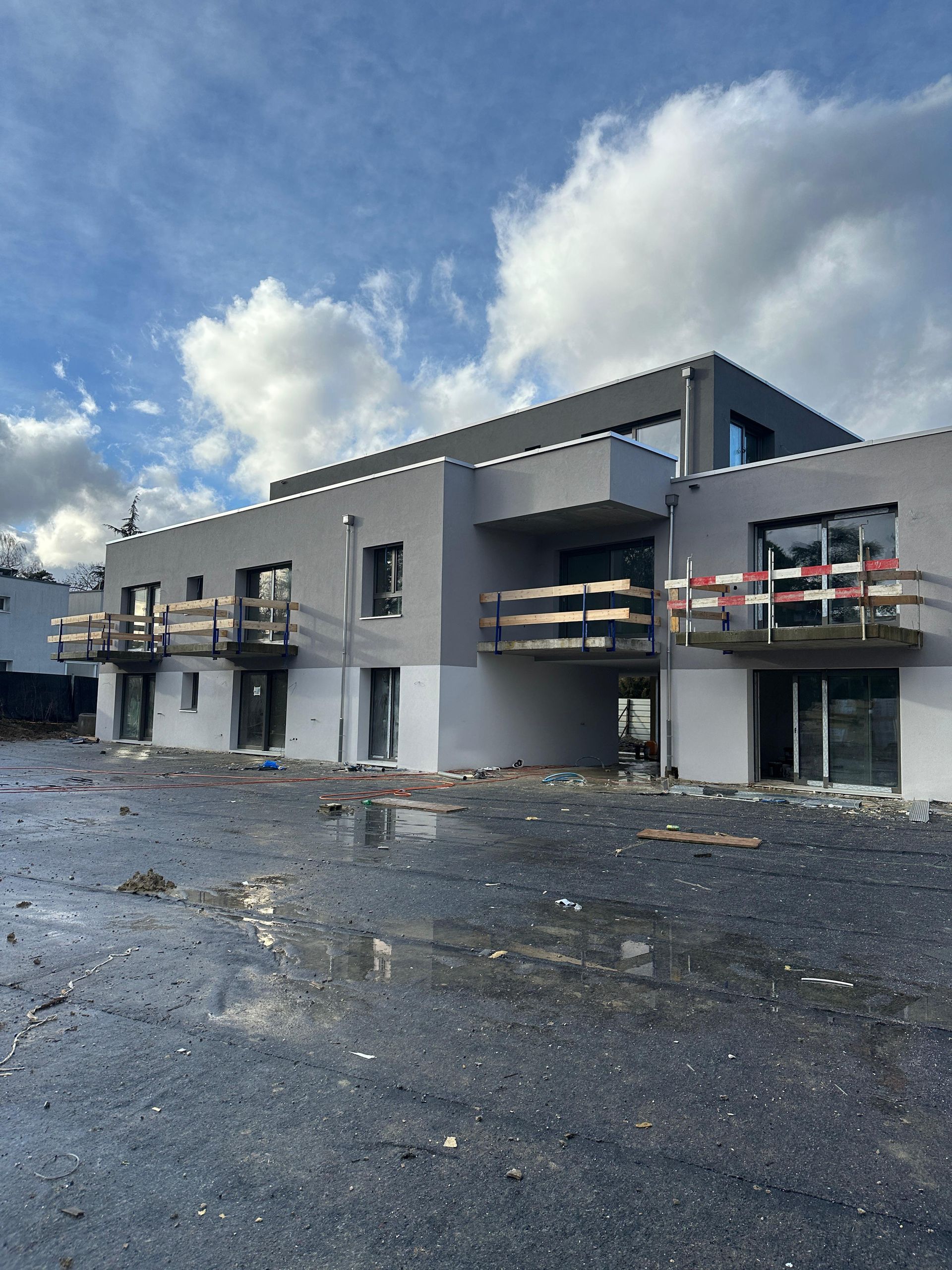Immeuble d'appartements moderne de deux étages avec façade en stuc gris, balcons en bois et un chantier de construction au premier plan.