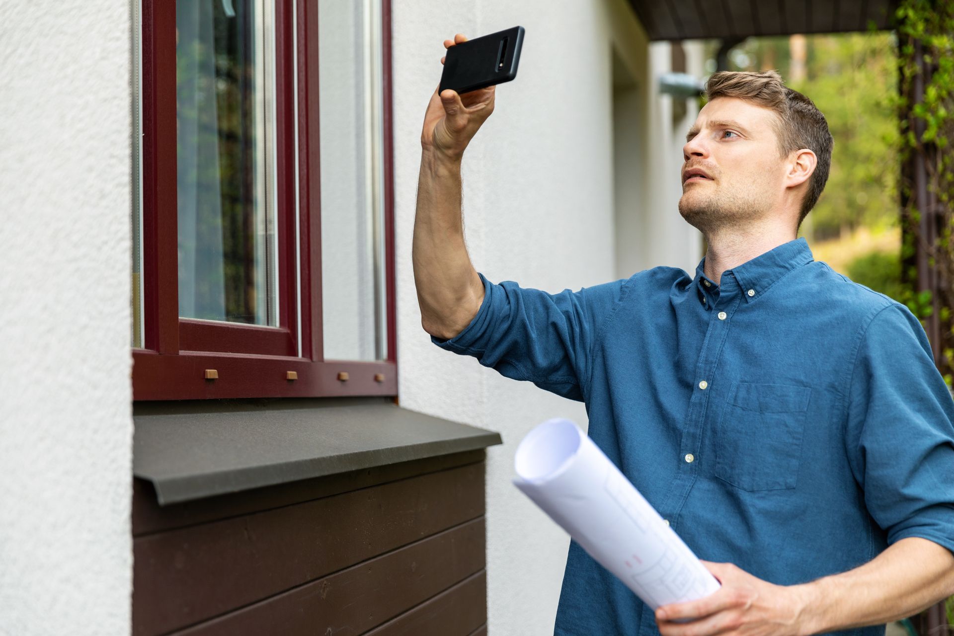 Man holding phone up to window, taking a photo, while holding a rolled blueprint in front of white building with brown window trim.