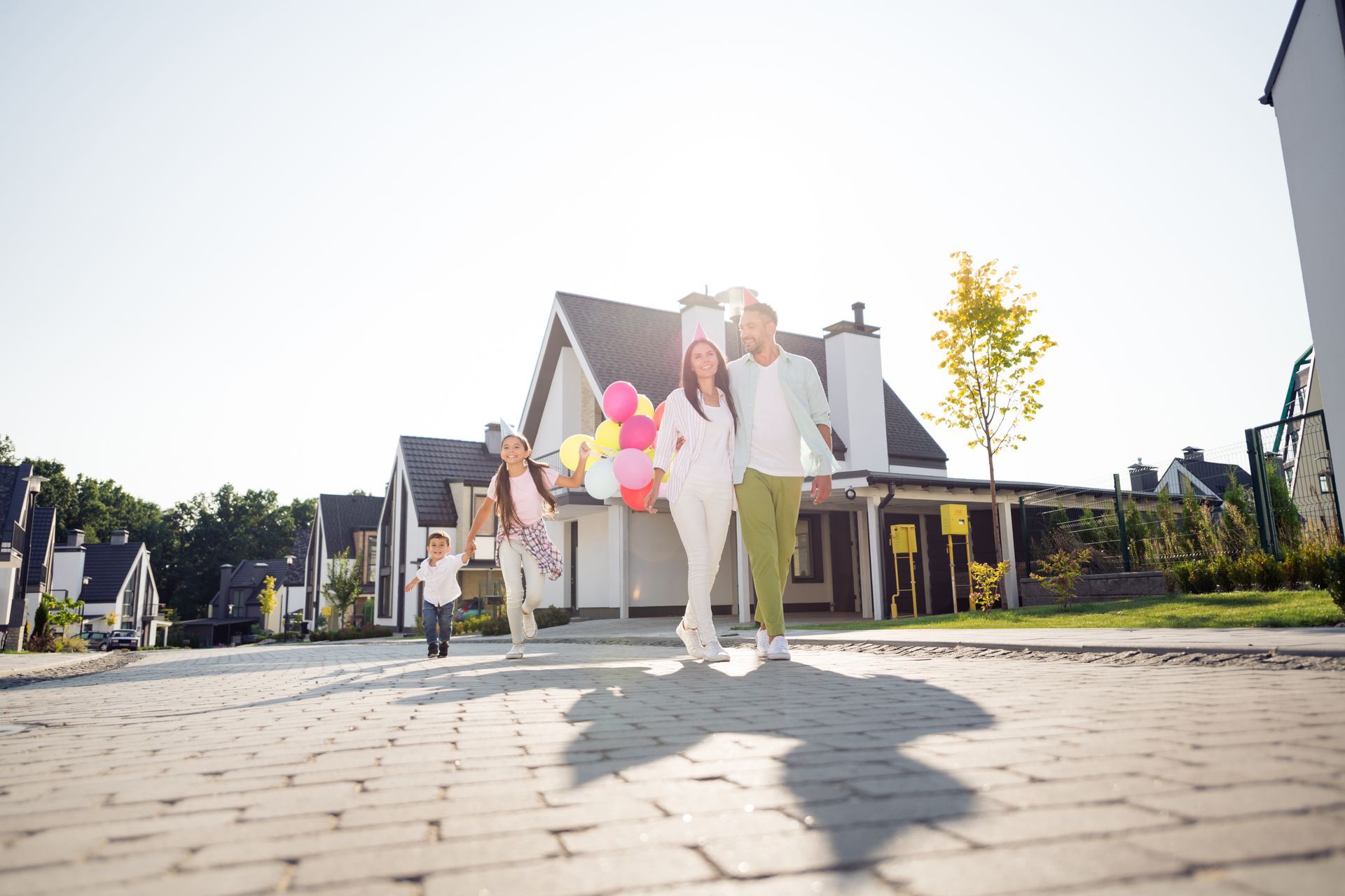 Family walking down a brick-paved street holding balloons near houses on a sunny day.