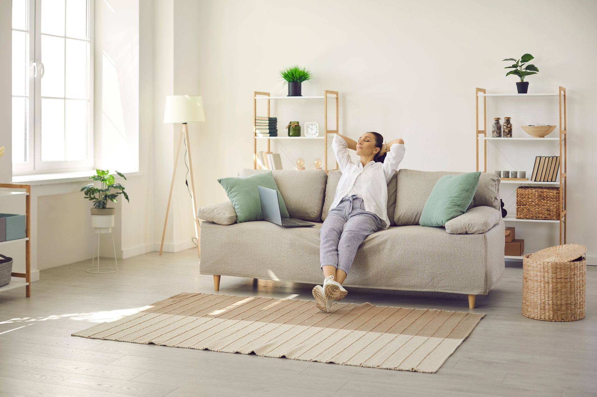 Woman relaxing on a sofa with laptop, arms behind head, in a bright living room.
