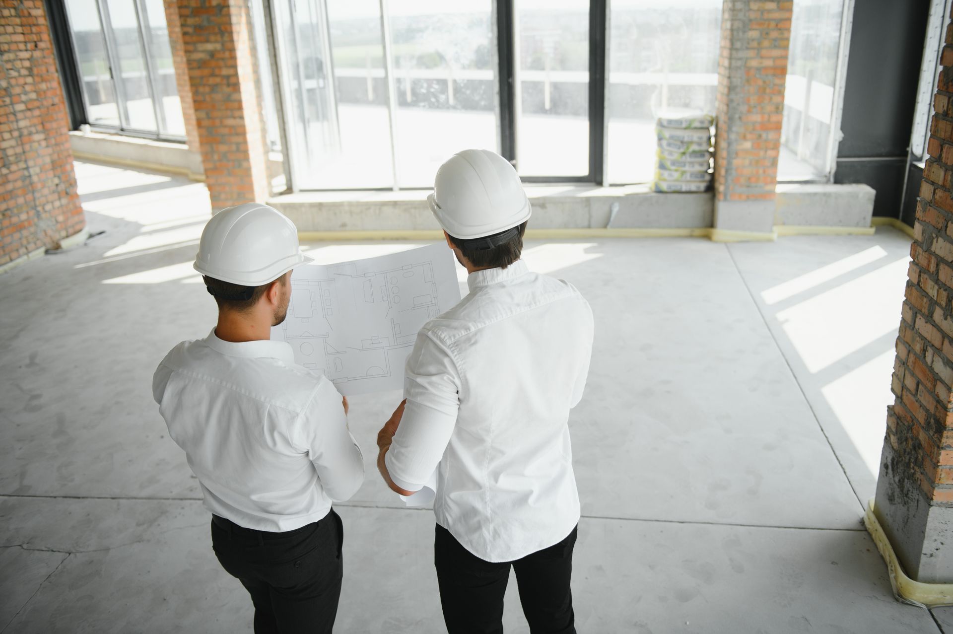 Two people in white hard hats and shirts, reviewing blueprints in an unfinished building.