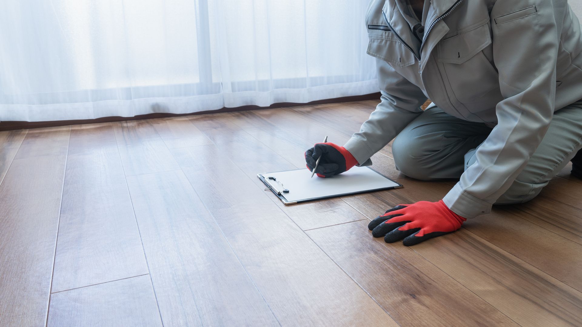 Person in work clothes kneeling on a hardwood floor, writing on a clipboard.