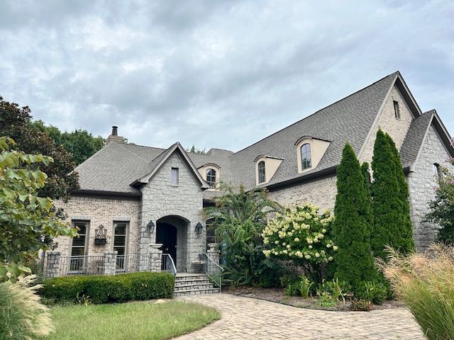 Stone house with gray roof, arched doorway, and manicured landscaping under a cloudy sky.