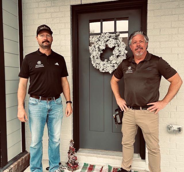 Two men in black shirts standing by a gray front door with a wreath. One wears a hat and jeans, the other tan pants.