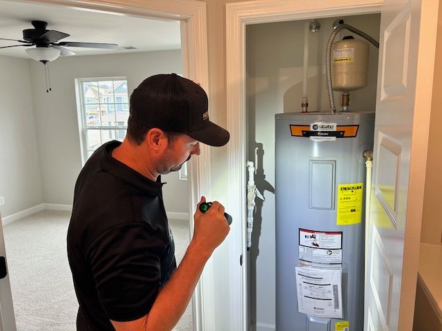 Man inspecting a water heater in a closet, holding a flashlight. Bedroom visible in the background.