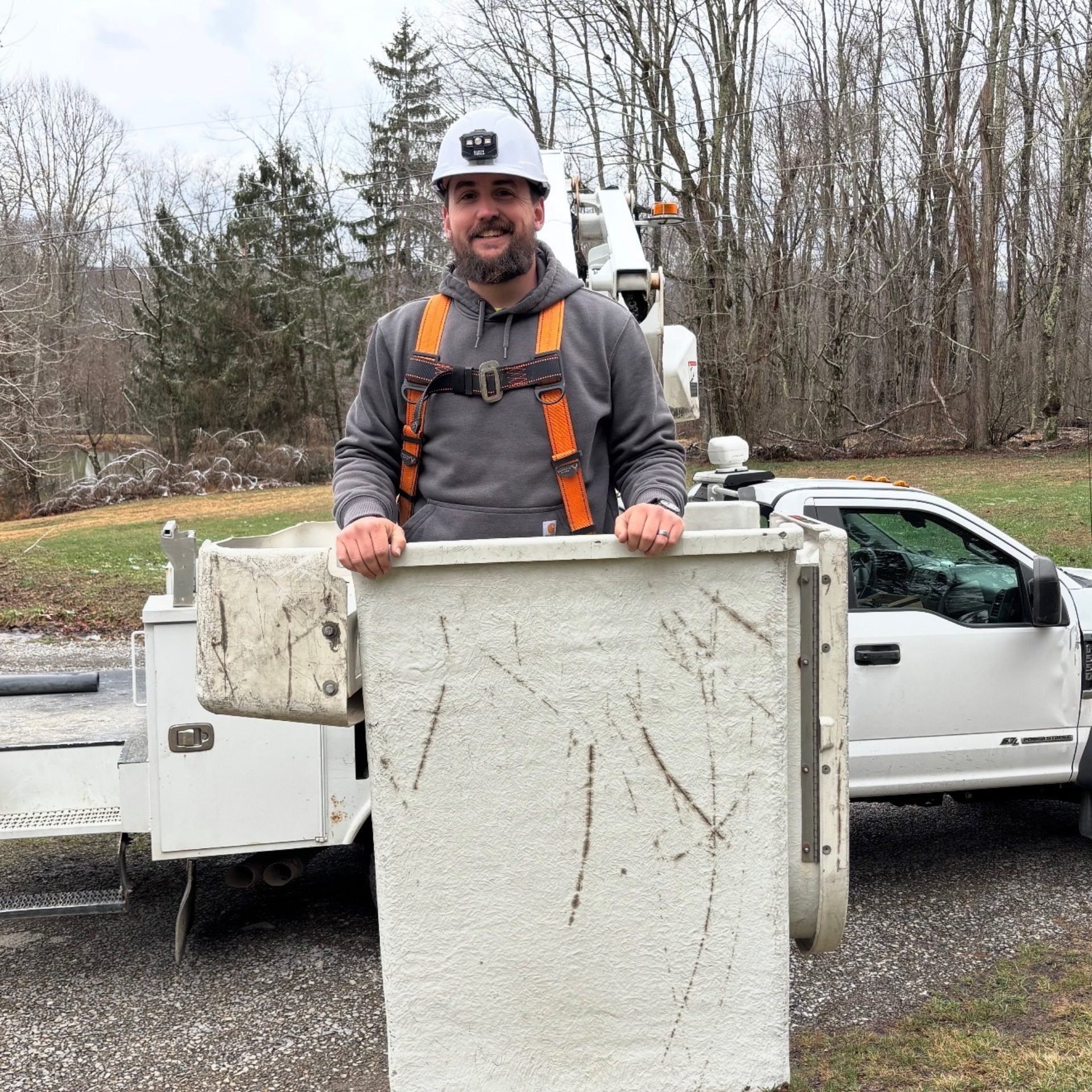 A person wearing a hard hat and safety harness stands in a white utility bucket attached to a truck parked in a field.