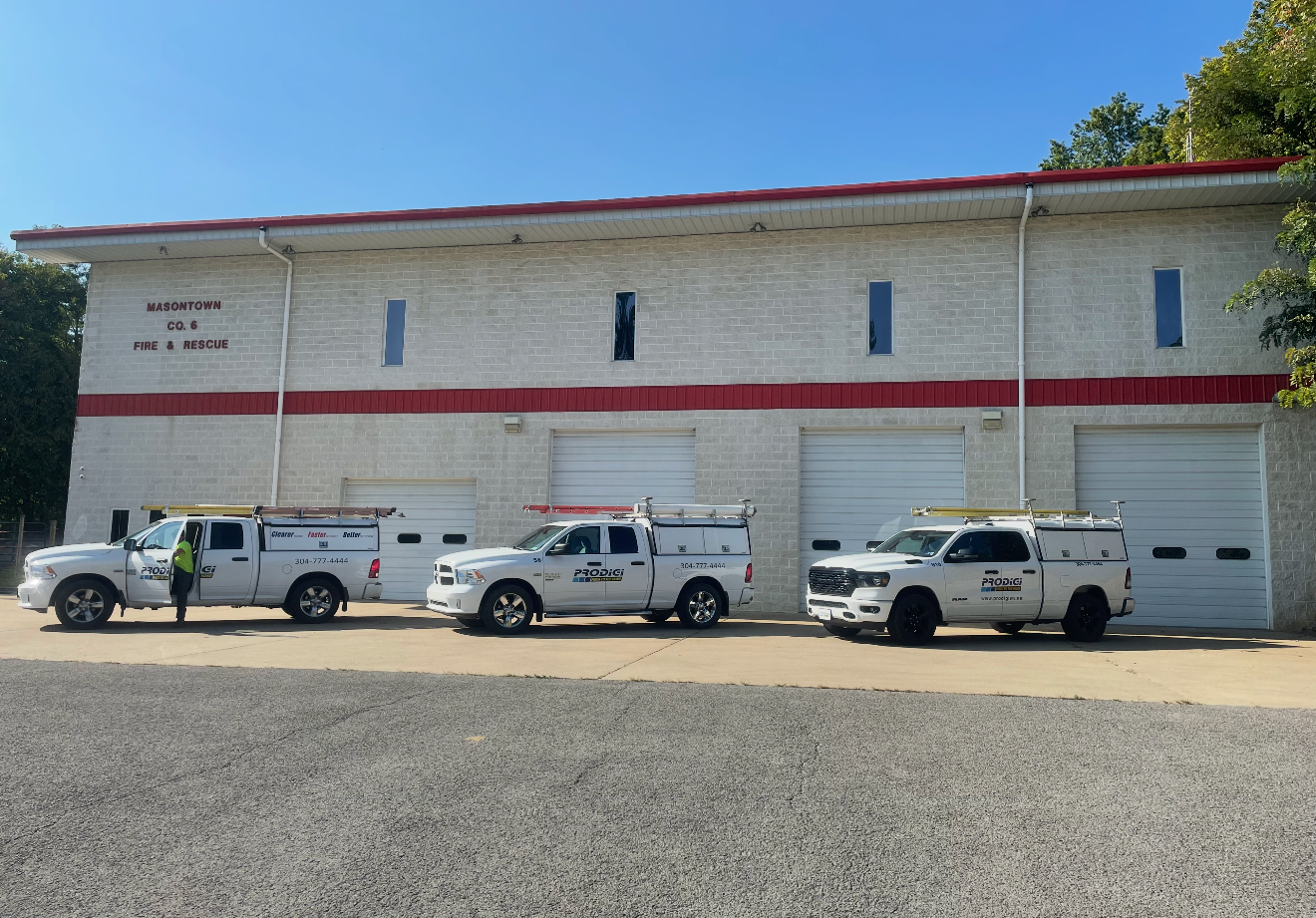 Three white service trucks parked in front of a white brick building with red trim and three garage bays on a gravel lot.