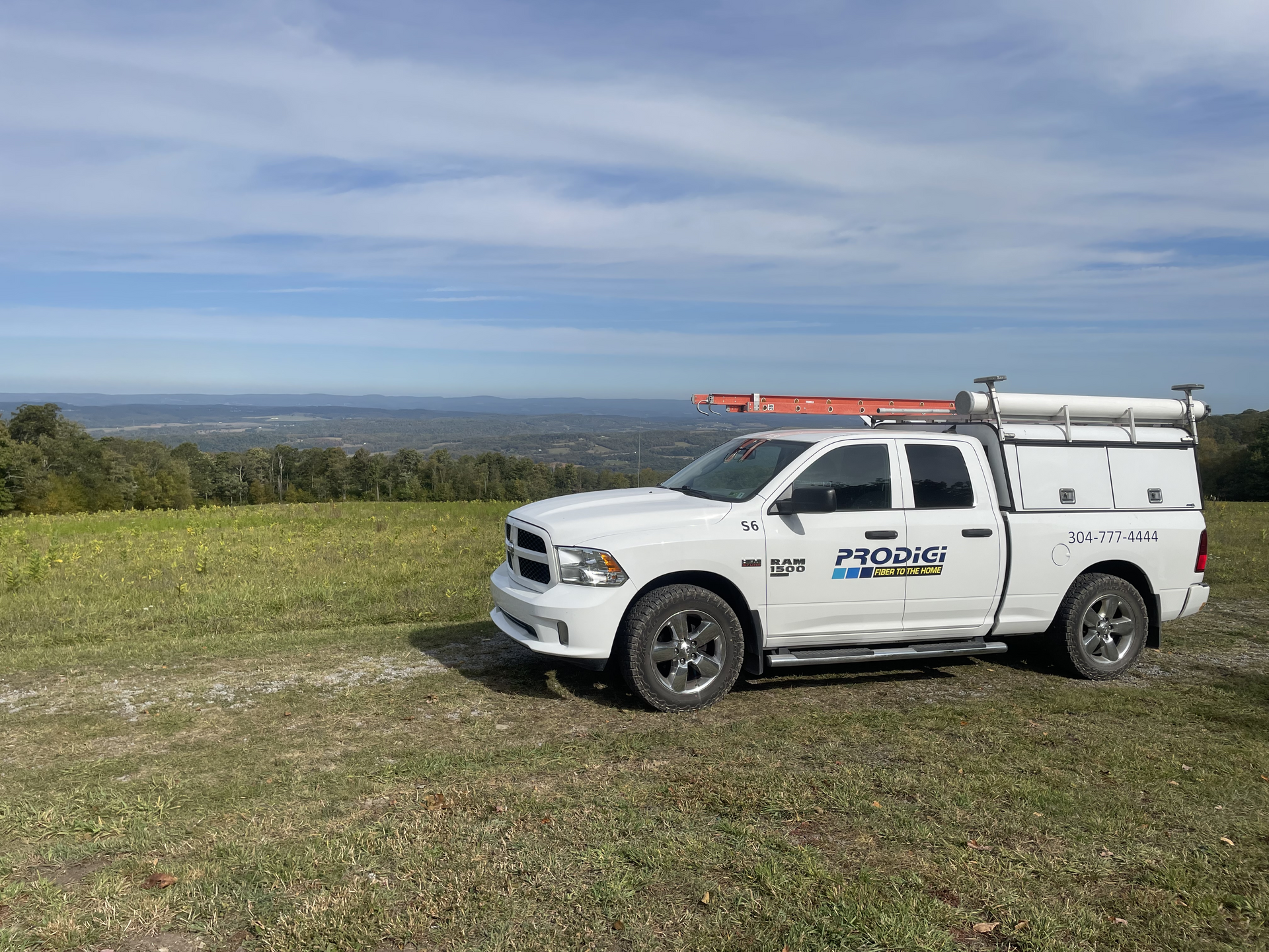 A white Ram pickup truck with a utility bed and a roof ladder rack parked on a grassy hill overlooking a valley.