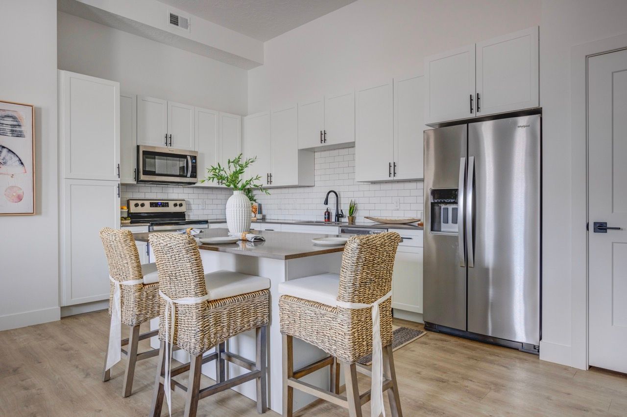 Modern white kitchen with island, stainless steel refrigerator, and woven bar stools.