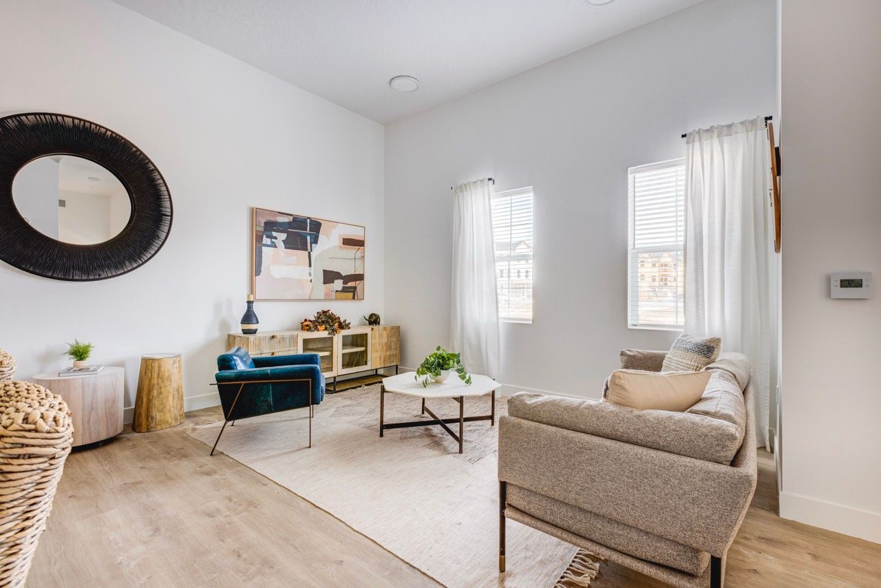 Bright living room with beige sofa, blue accent chair, round woven mirror, and two windows with white curtains.