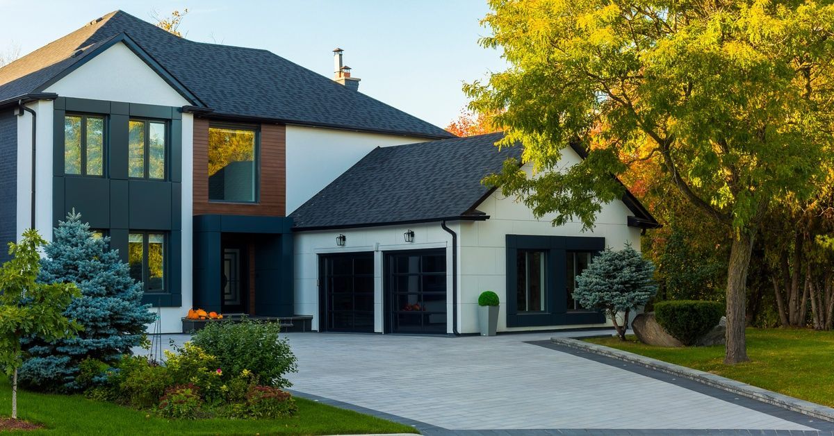 A modern white home contains dark trim, black garage doors, a wide driveway, and green trees around 
