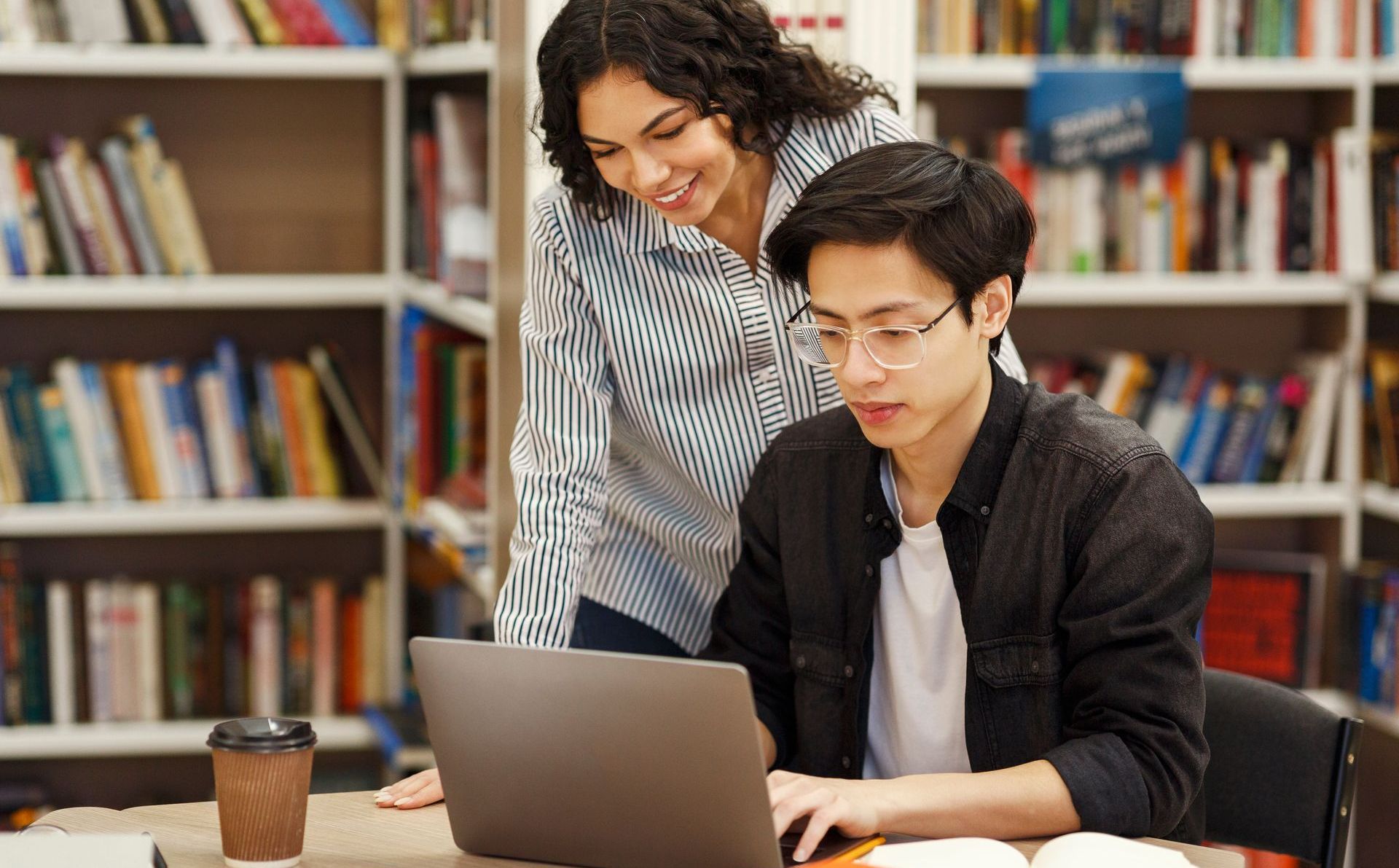 a woman is standing next to a man using a laptop computer in a library .