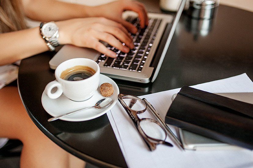 A woman is typing on a laptop computer while sitting at a table with a cup of coffee.