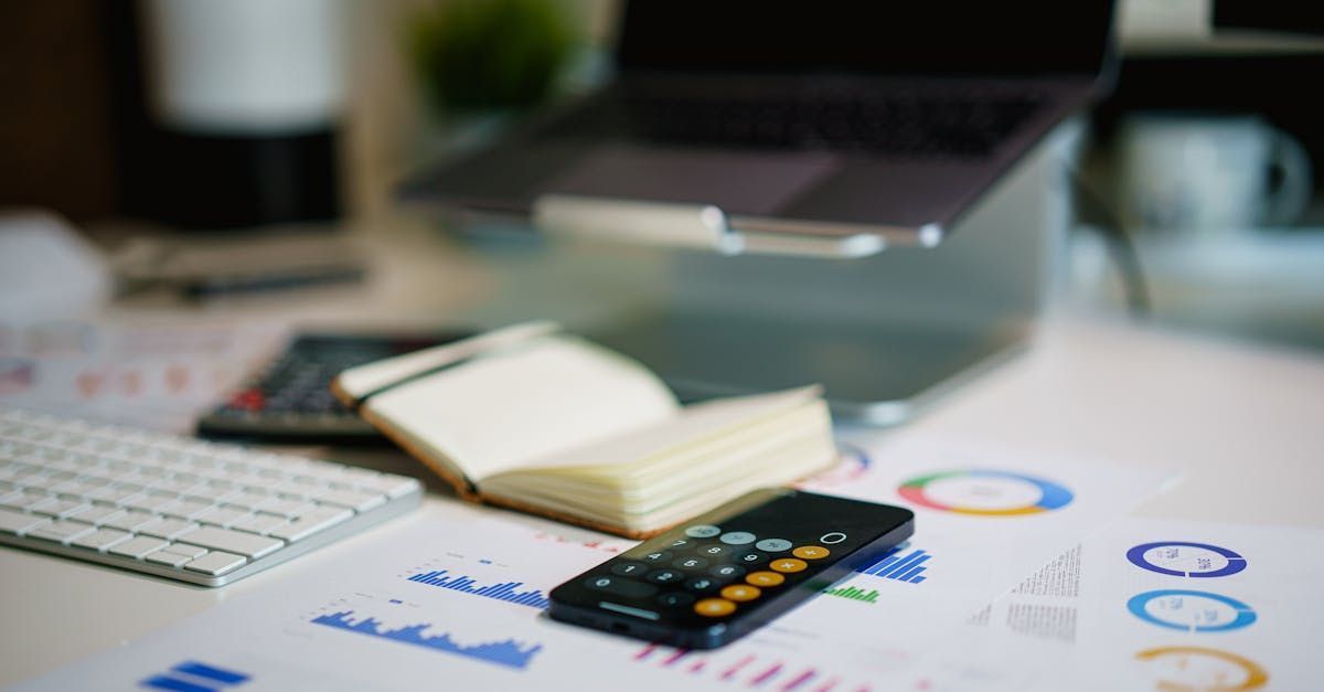 A book , calculator , cell phone , keyboard and laptop are on a desk.