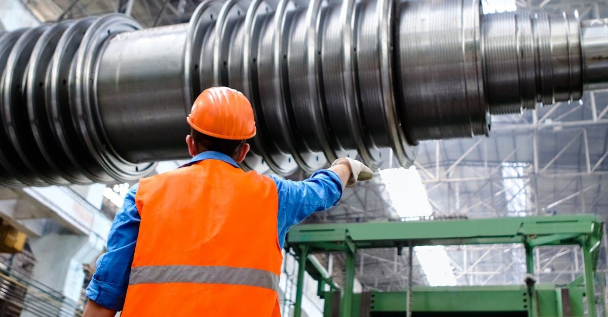 A man in an orange vest and hard hat is working on a large metal object in a factory.
