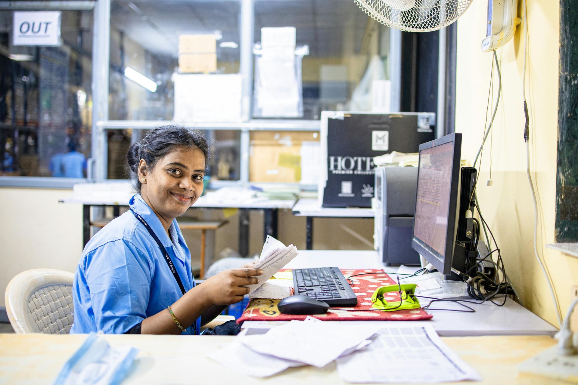 A woman is sitting at a desk in front of a computer holding a piece of paper.