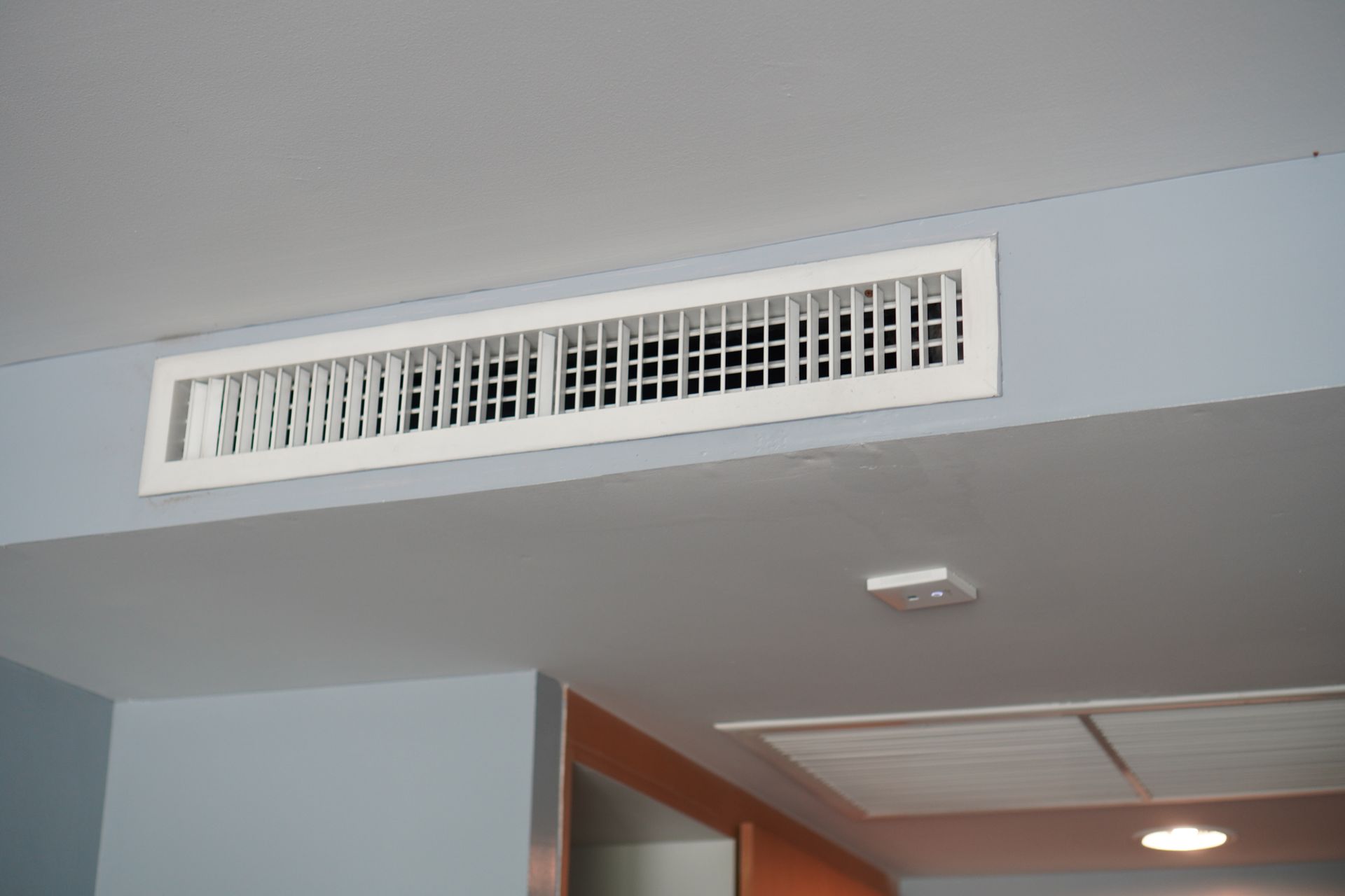 Hand holding AC remote, aiming at wall-mounted air conditioner. Room interior with lamp, curtains, and peach-colored wall.