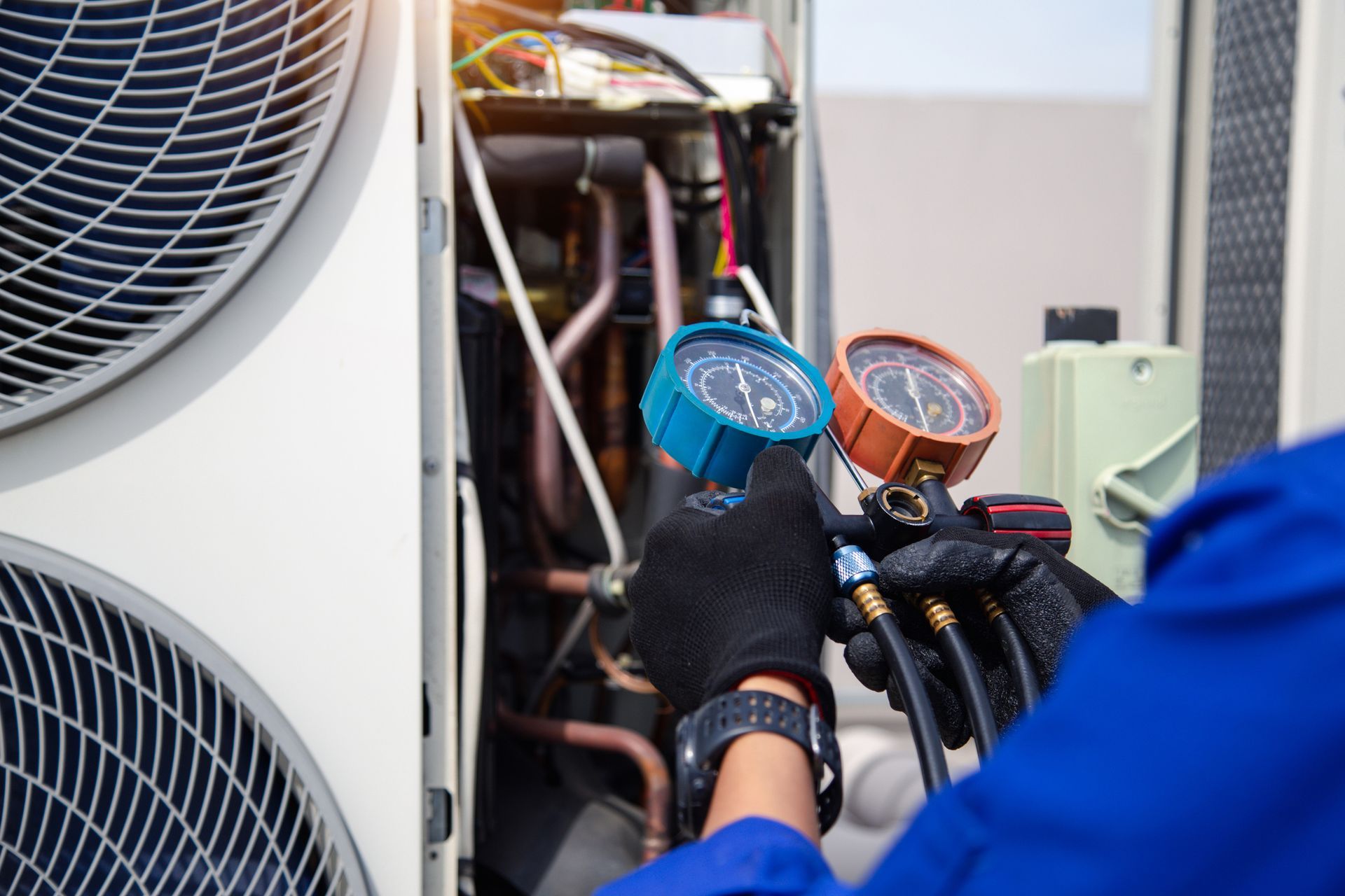 HVAC technician in blue overalls using gauges to service an outdoor air conditioning unit.