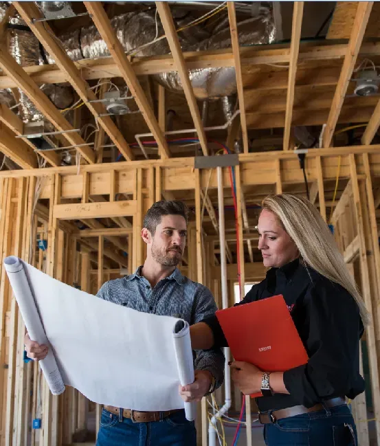 Man and woman reviewing blueprints at a construction site. They stand inside a wooden frame.