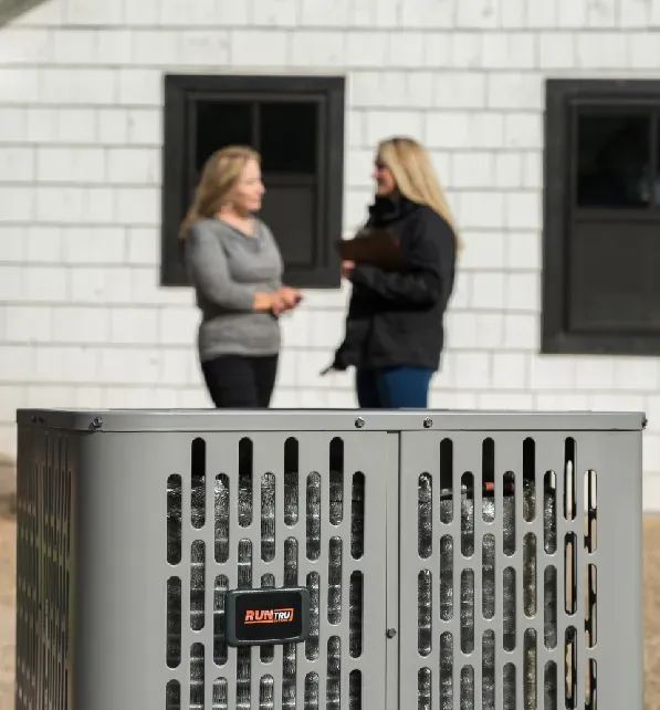 Two women discussing near an AC unit in front of a white house with black windows.