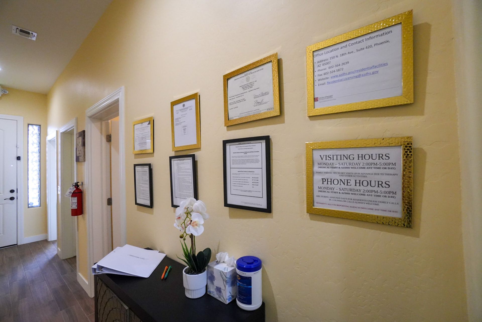 Hallway with framed documents on the wall. A black table with an orchid, tissues, and a bottle sits below.