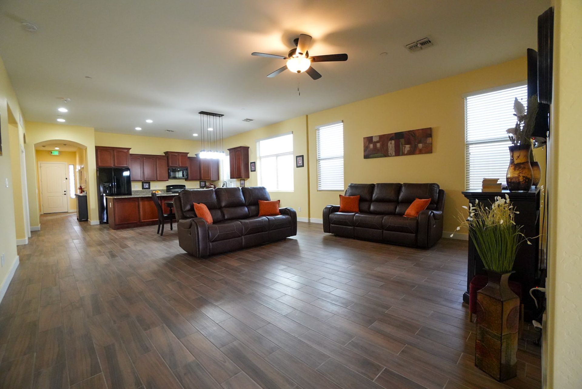 Spacious living area with brown leather sofas, open to a kitchen with dark cabinetry and wood-look flooring.