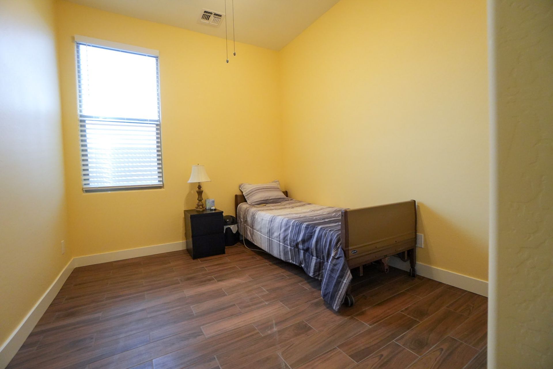 Small bedroom with yellow walls, a single bed, a nightstand, and a window with blinds. Wooden floor.