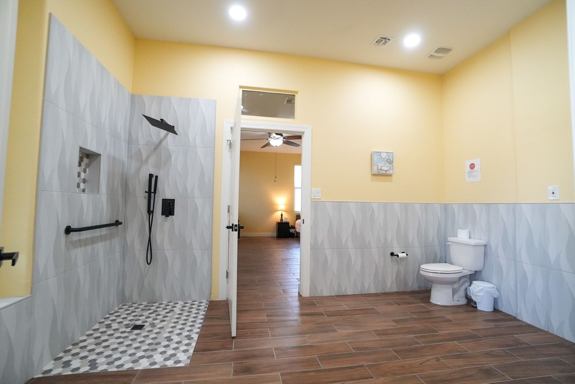 Accessible bathroom with a roll-in shower, toilet, and doorway to a living area; light yellow walls, wood-look floor.
