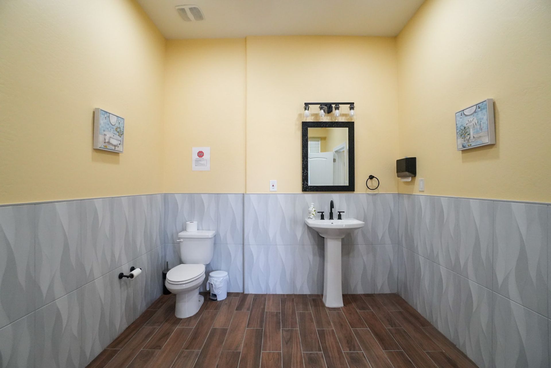 A public restroom with beige walls, gray tile wainscot, and wood-look flooring; includes a toilet, sink, and mirror.